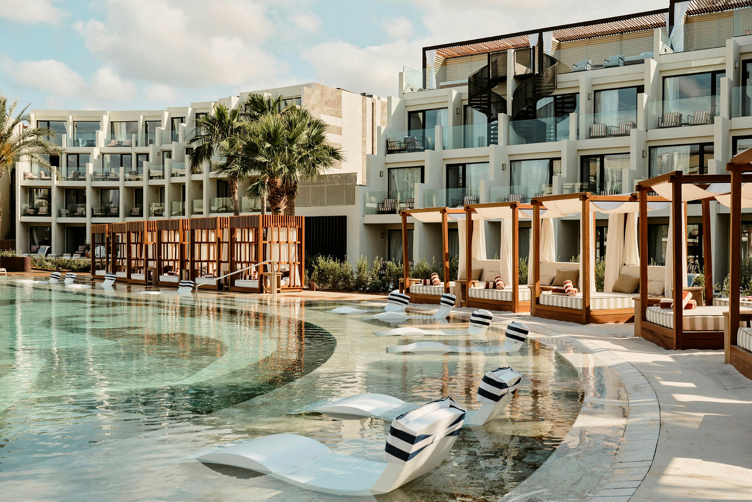 En hotelpool med liggestole i poolen og cabanas ved siden af. Bygningen har altaner med glasrækværk. (AI-genereret tekst)