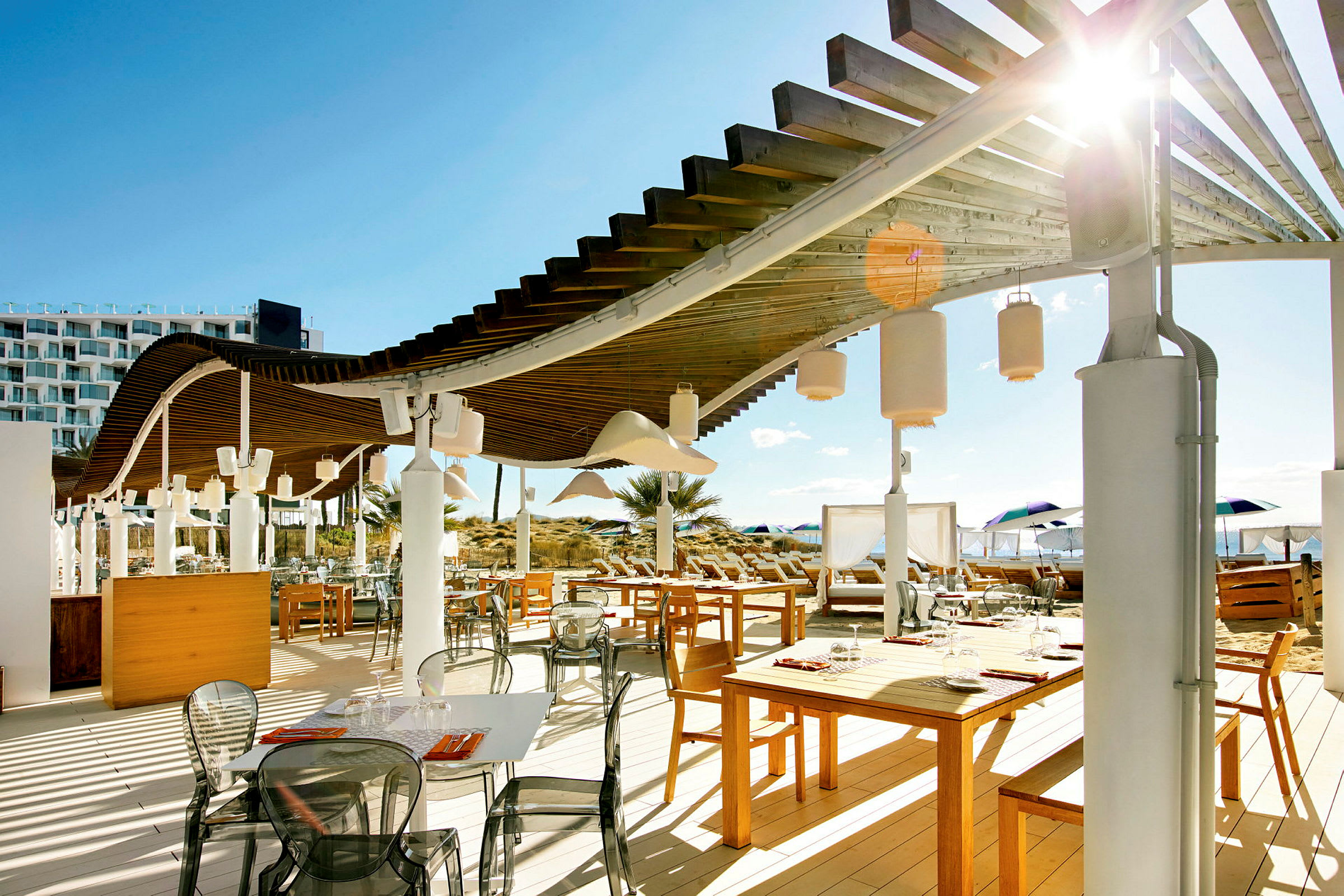 En udendørsrestaurant med træborde, stole og en pergola over. I baggrunden ses strand og parasoller. (AI-genereret tekst)