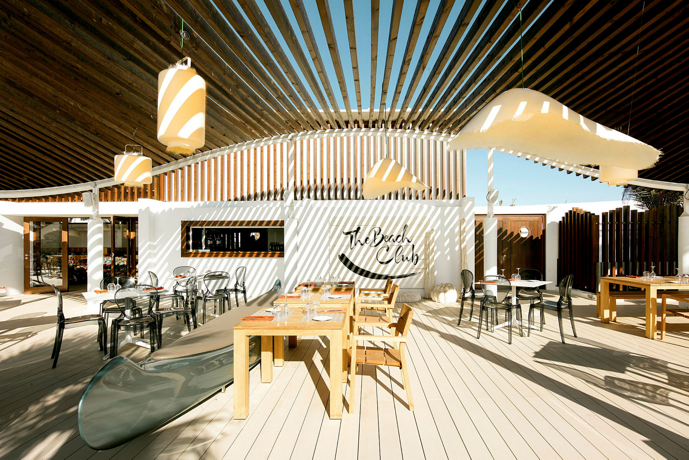 Udendørsområde med borde og stole samt dekoration med The Beach Club-skiltet. (AI-genereret tekst)