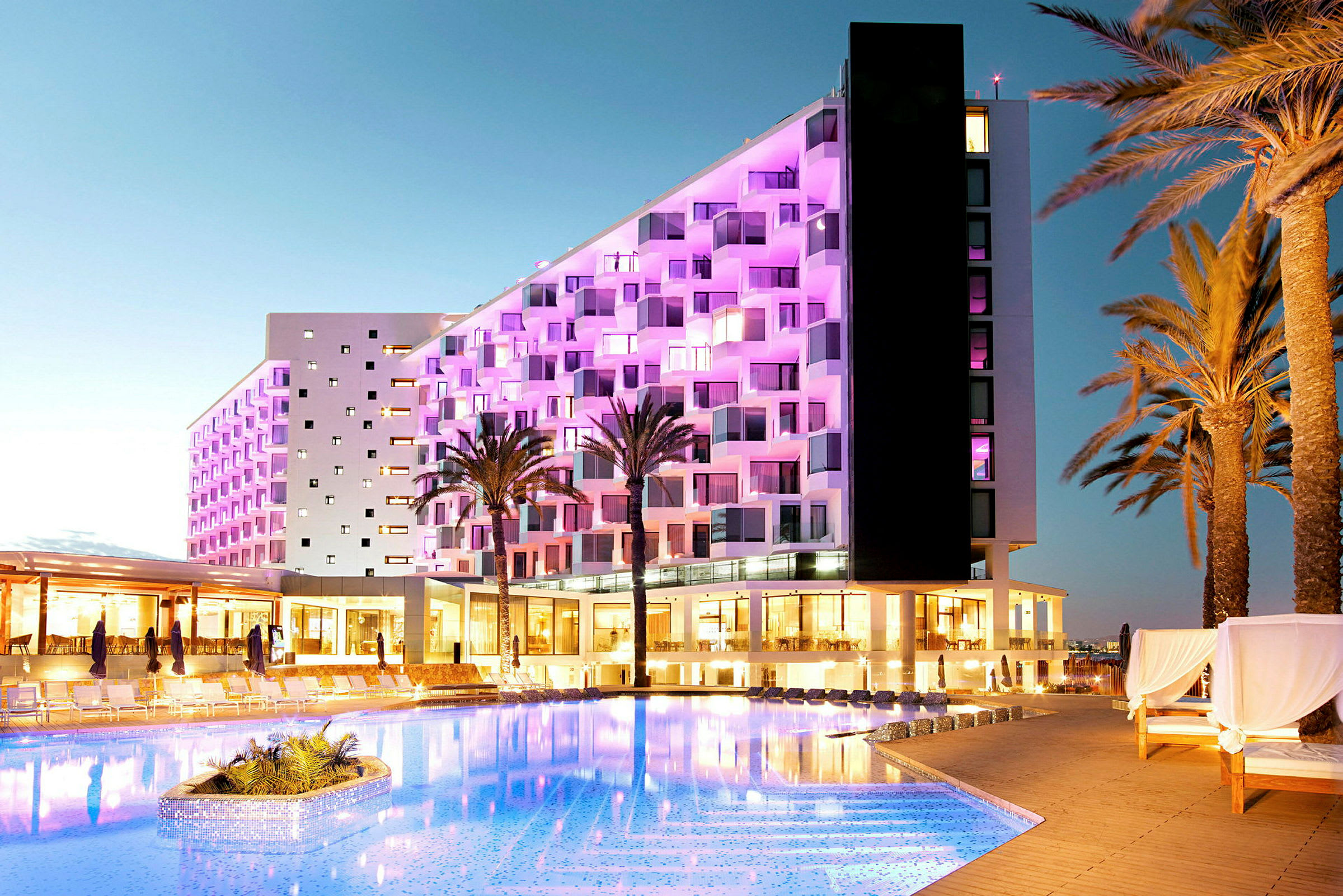 Et hotel med facade oplyst af pink lys ved siden af en pool, omgivet af palmetræer. (AI-genereret tekst)