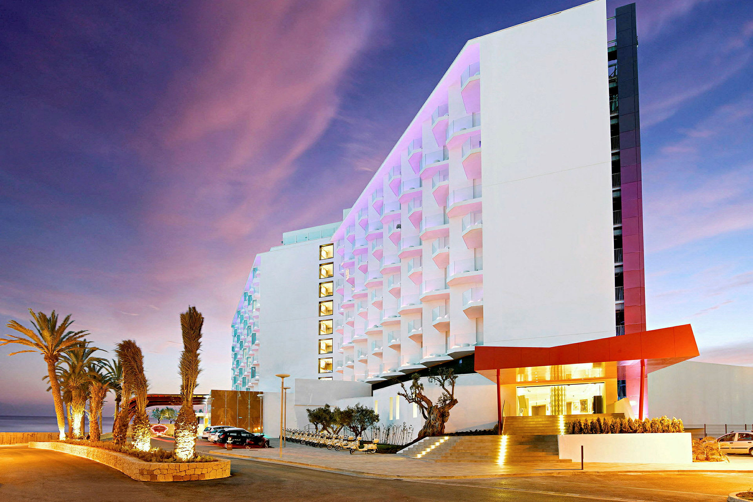 Moderne hotel med oplyste palmer ved hovedindgangen og en farverig facade. (AI-genereret tekst)