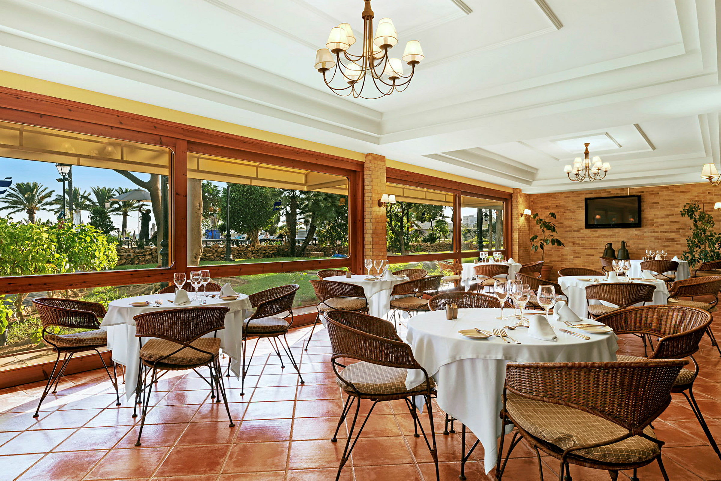 En elegant restaurant med dækkede borde og fletstole. Store vinduer giver udsigt til en have. (AI-genereret tekst)