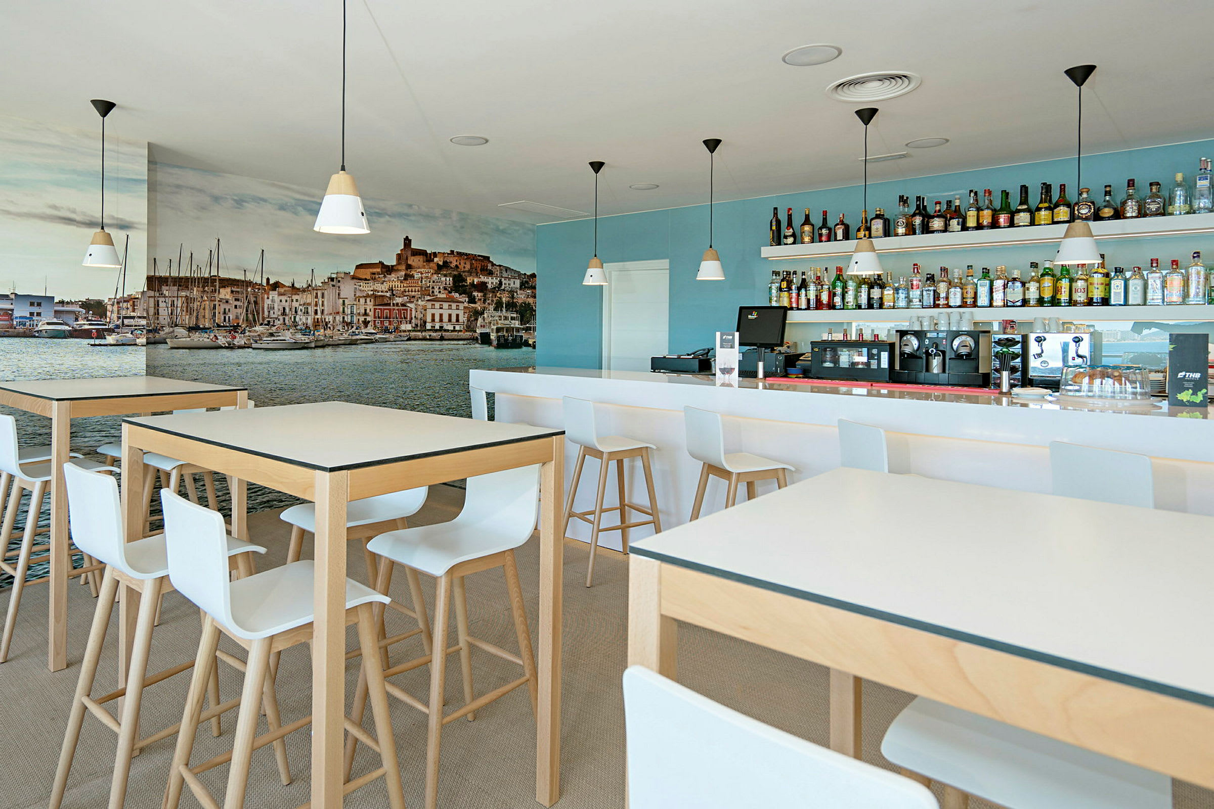 En moderne café med høje borde og stole, en bar med drikkevarer i baggrunden og et stort vægmaleri af en havneby. (AI-genereret tekst)