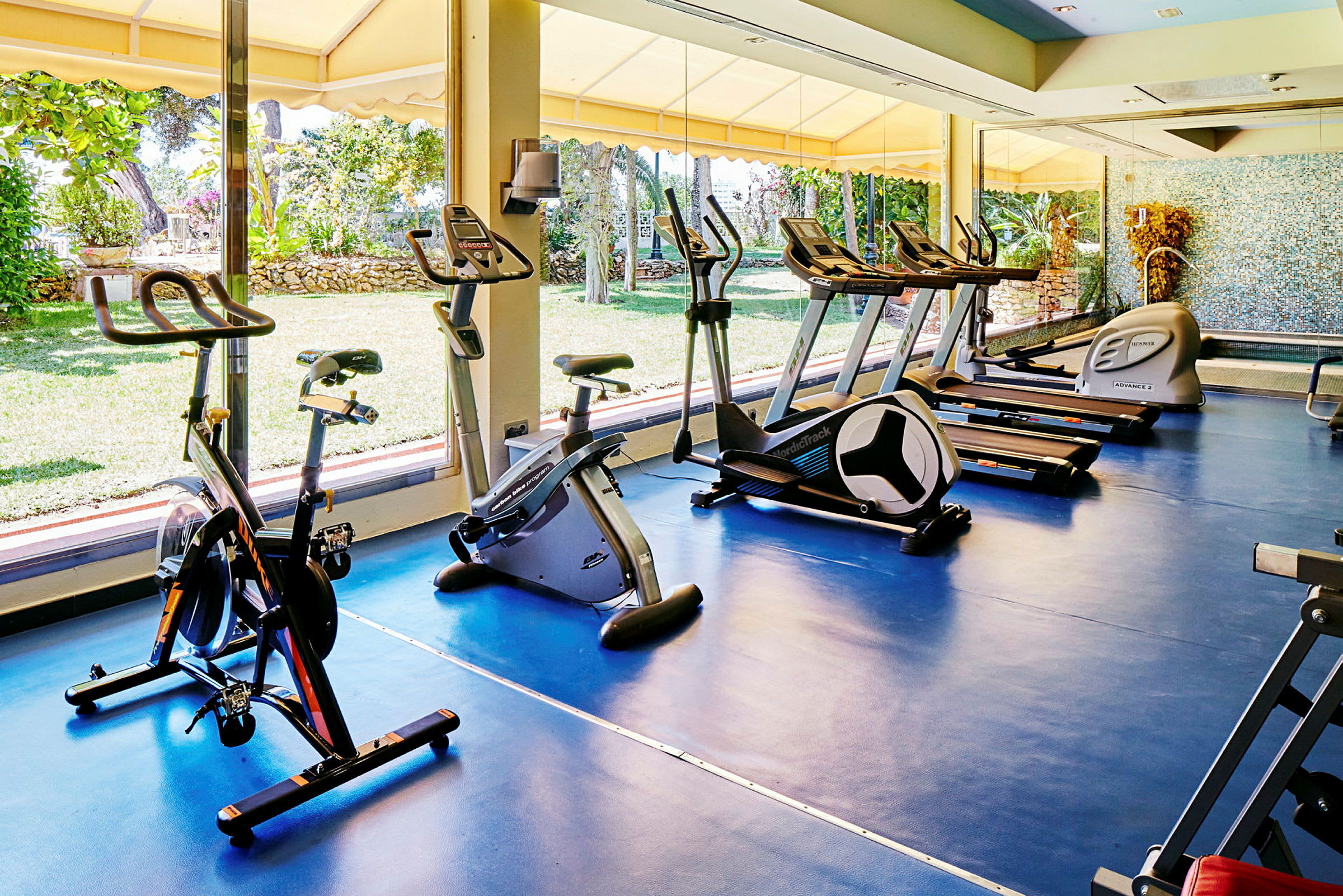 Et fitnesscenter med flere træningsmaskiner, herunder motionscykler og løbebånd, placeret ved et stort vindue med udsigt til en have. (AI-genereret tekst)