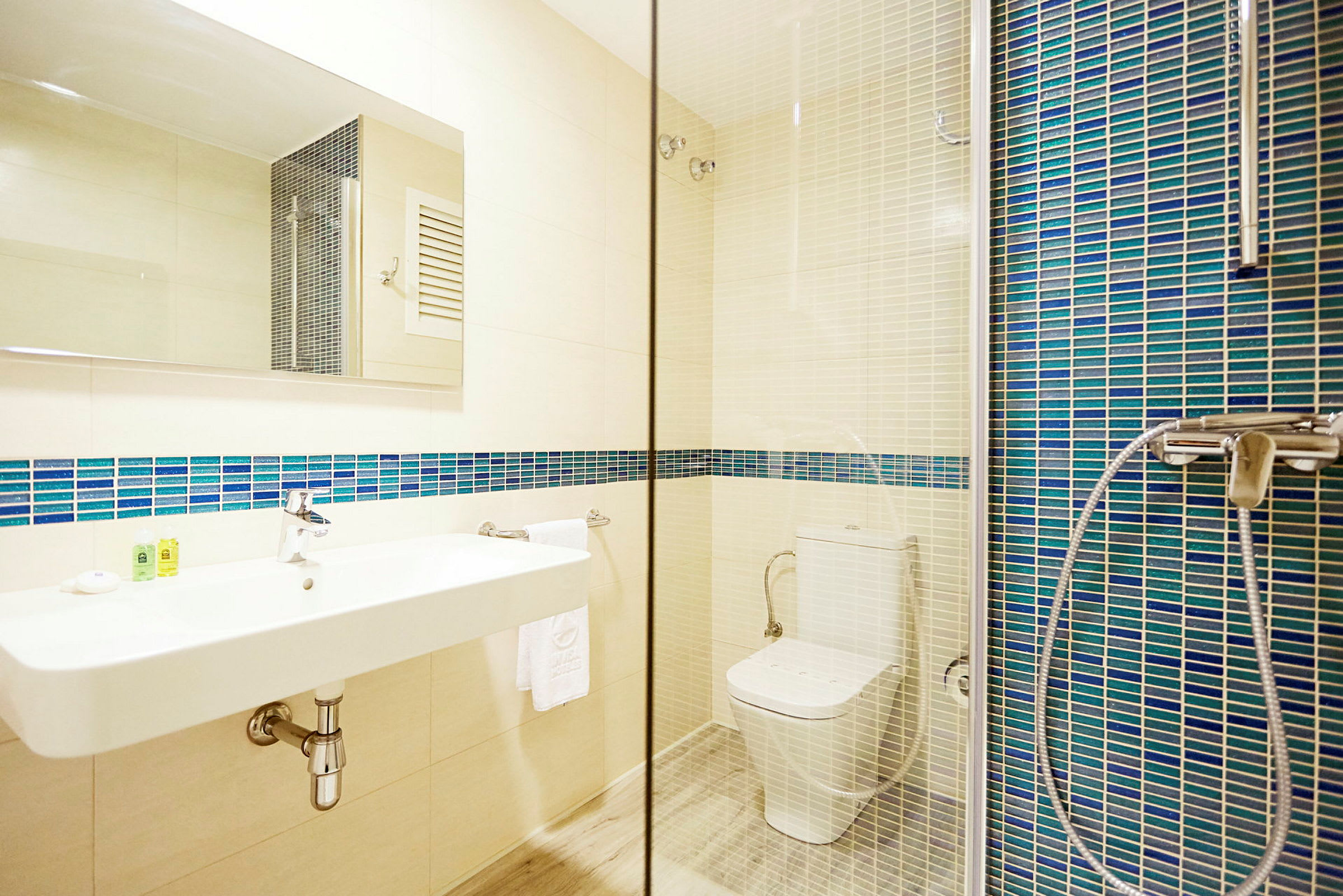 Et badeværelse med en hvid håndvask, et toilet og en bruser samt blå mosaikfliser på væggen. (AI-genereret tekst)
