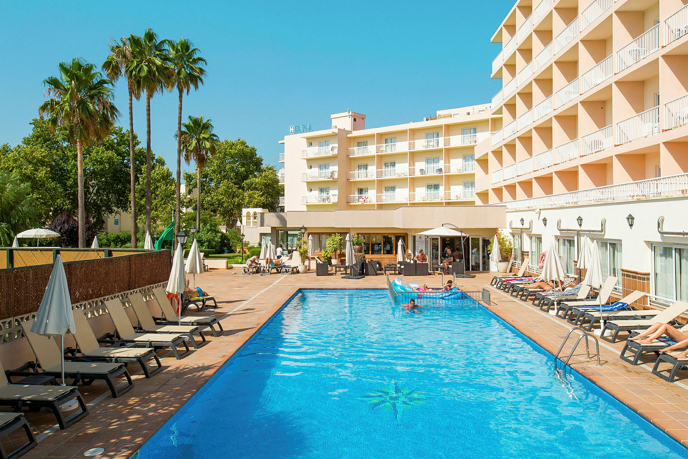 En hotelbygning ses ved siden af en udendørs pool med liggestole og parasoller omkring. (AI-genereret tekst)