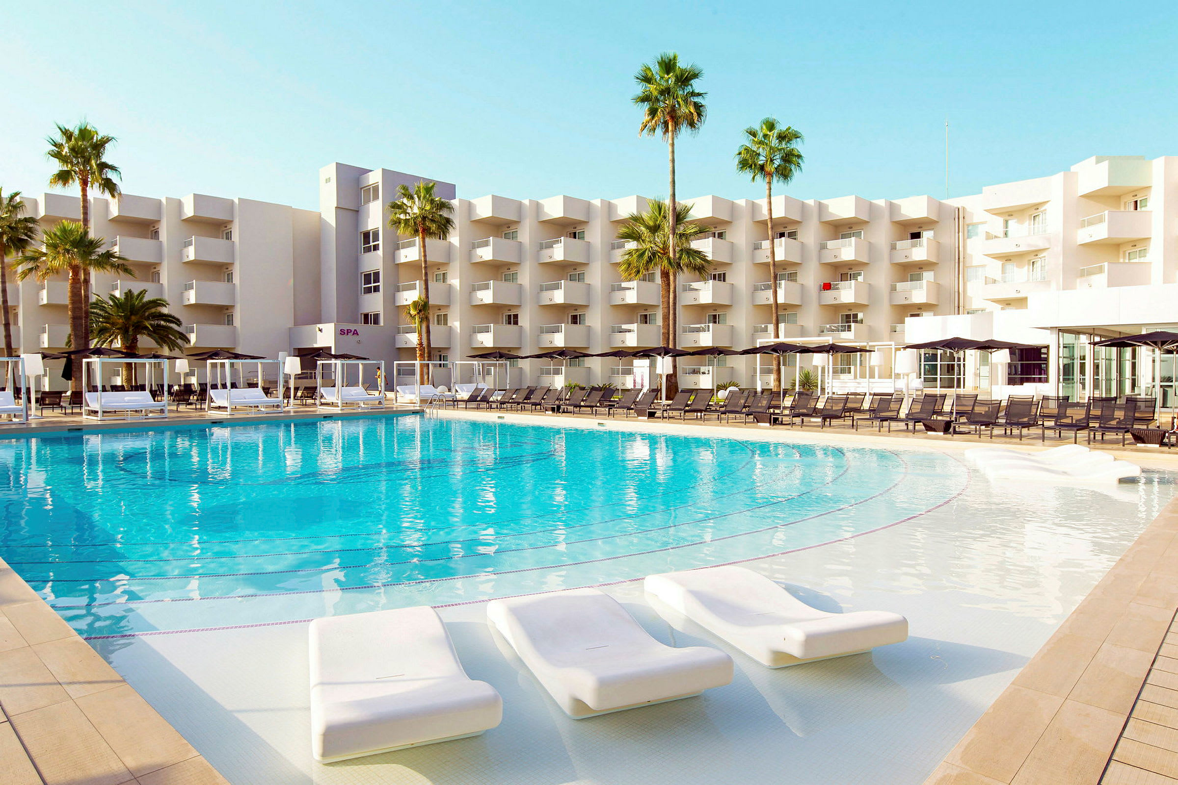 Stor swimmingpool omgivet af liggestole foran en moderne hotelbygning og palmer. (AI-genereret tekst)