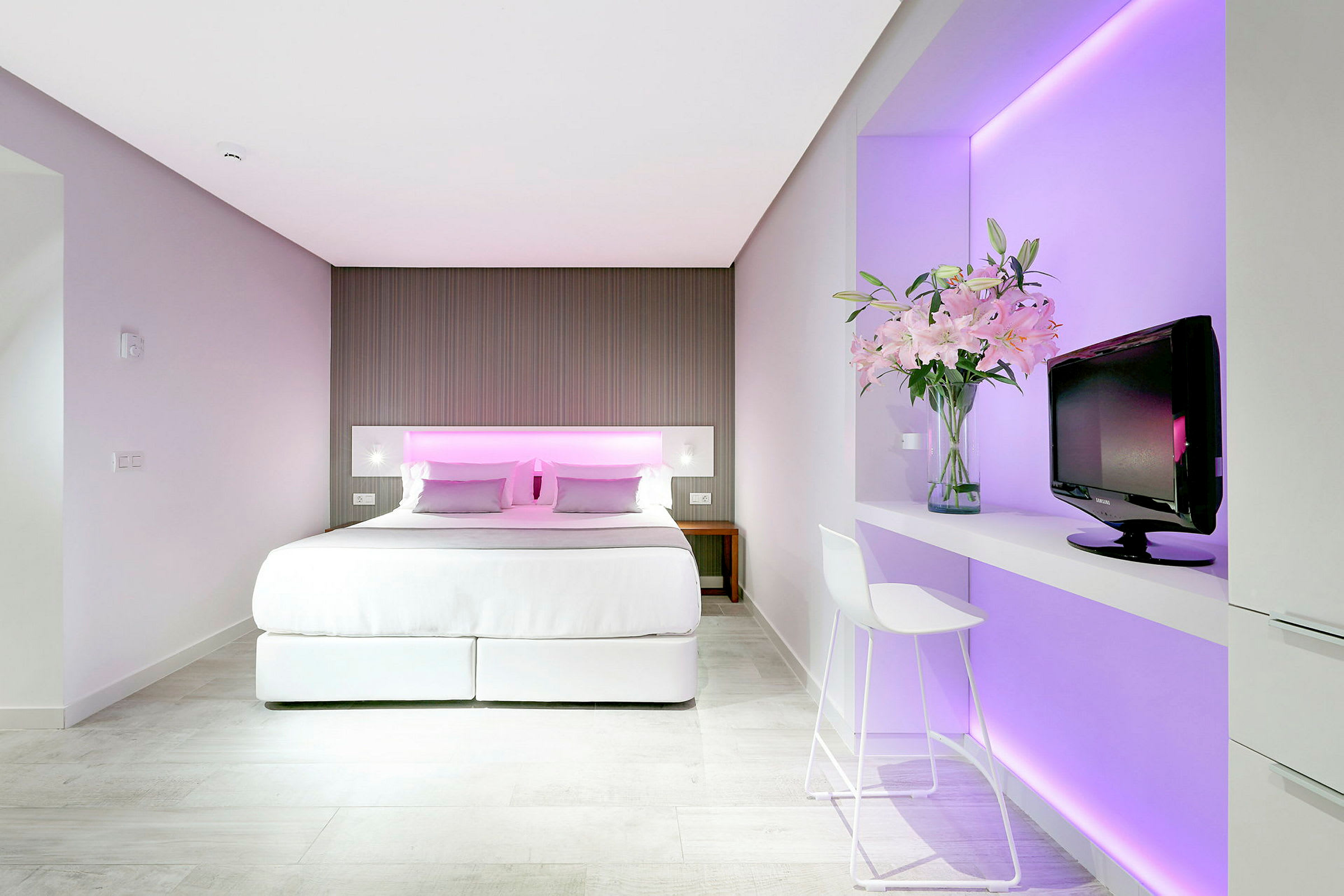Hotelværelse med dobbeltseng, minimalistisk indretning og pink belysning bag sengegavlen. Et tv og en vase med blomster står på en hylde ved siden af. (AI-genereret tekst)