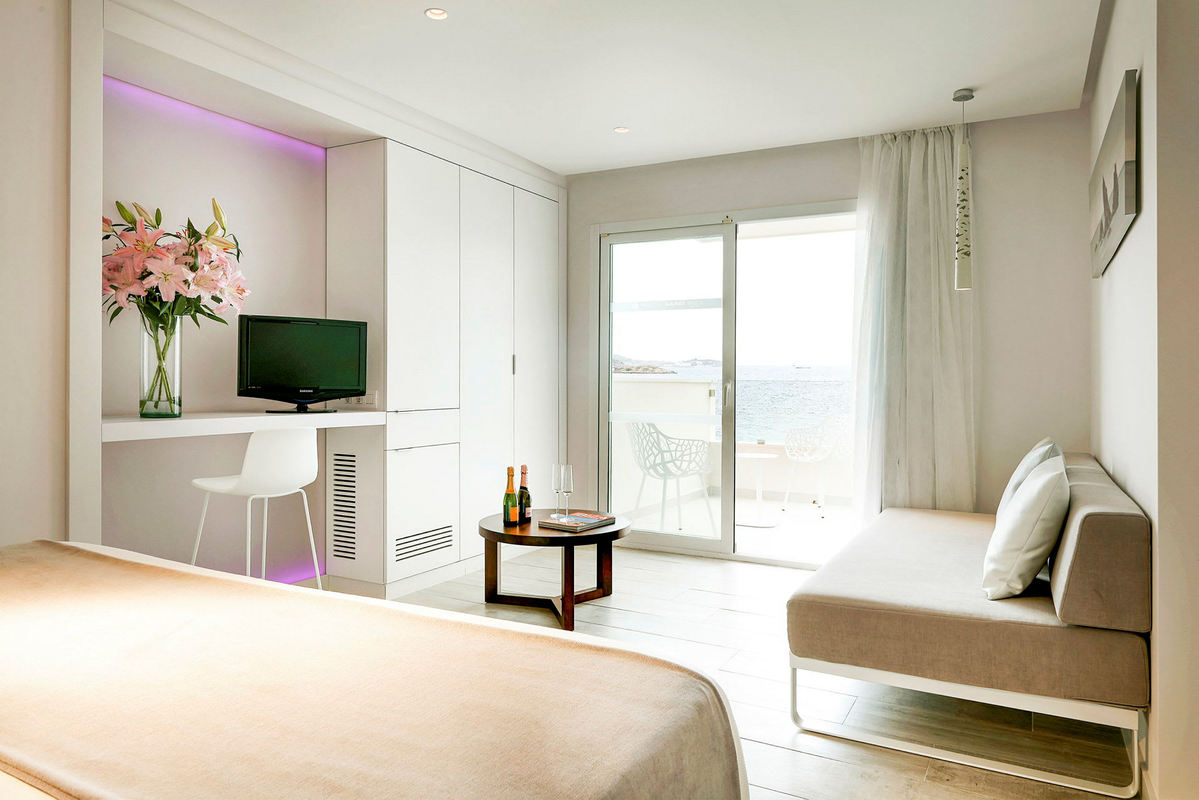 Et lyst hotelværelse med en seng, en sofa, et bord og en balkon med udsigt over havet. (AI-genereret tekst)