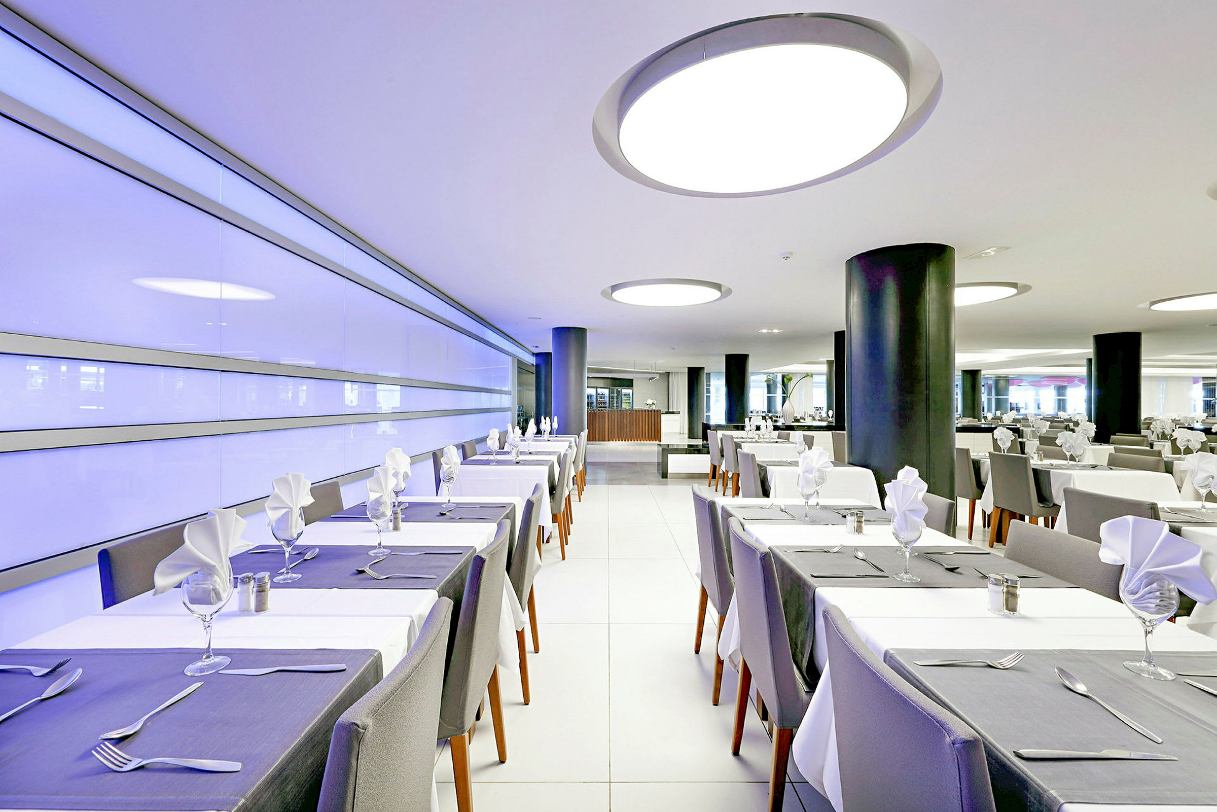 En moderne restaurant med dækkede borde og hvide stole. Interiøret inkluderer moderne lamper og en lys atmosfære. (AI-genereret tekst)