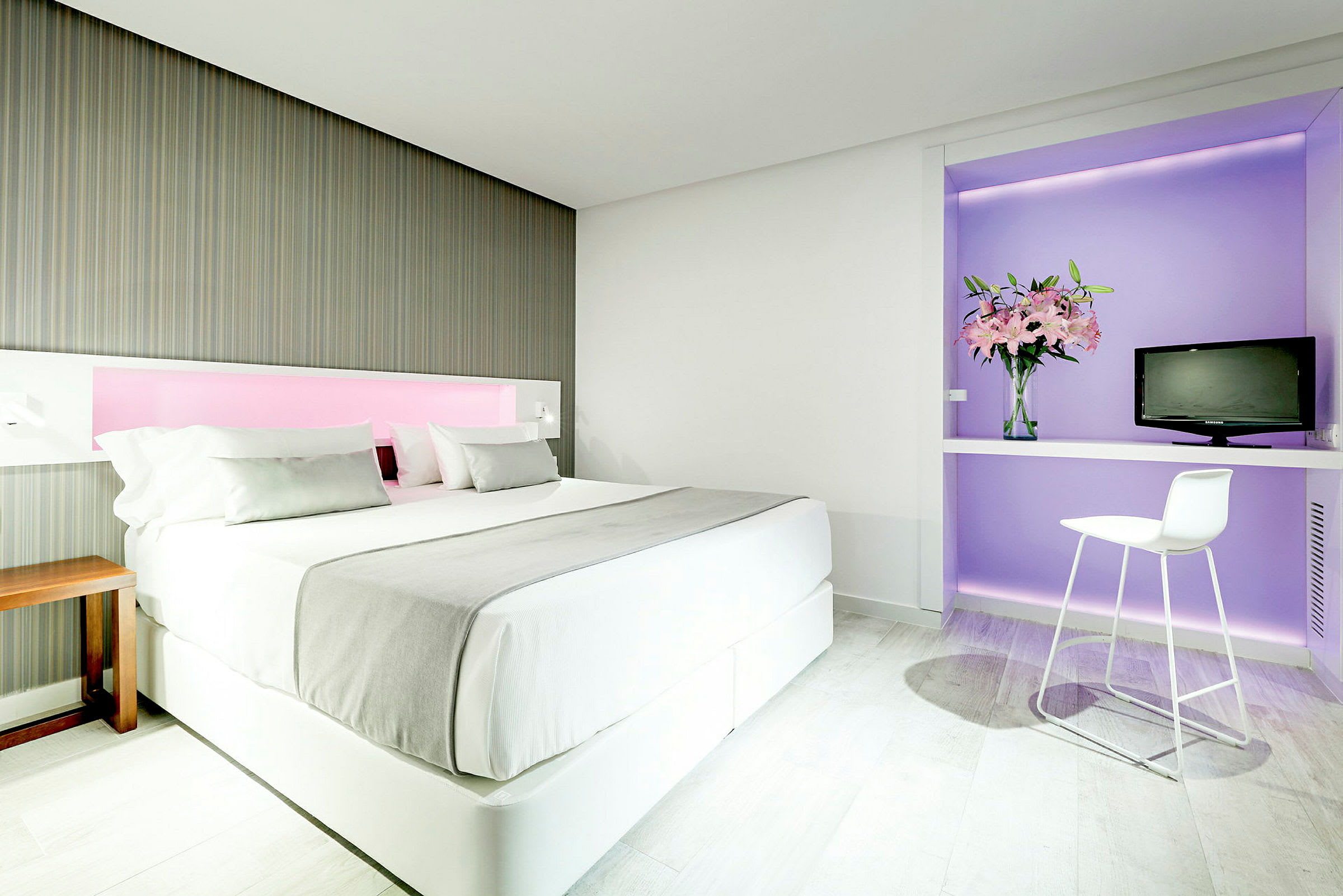 Et moderne hotelværelse med en stor seng, dekoreret i lyse farver. Til højre er der et skrivebord med en stol, en vase med blomster og et lille TV. (AI-genereret tekst)