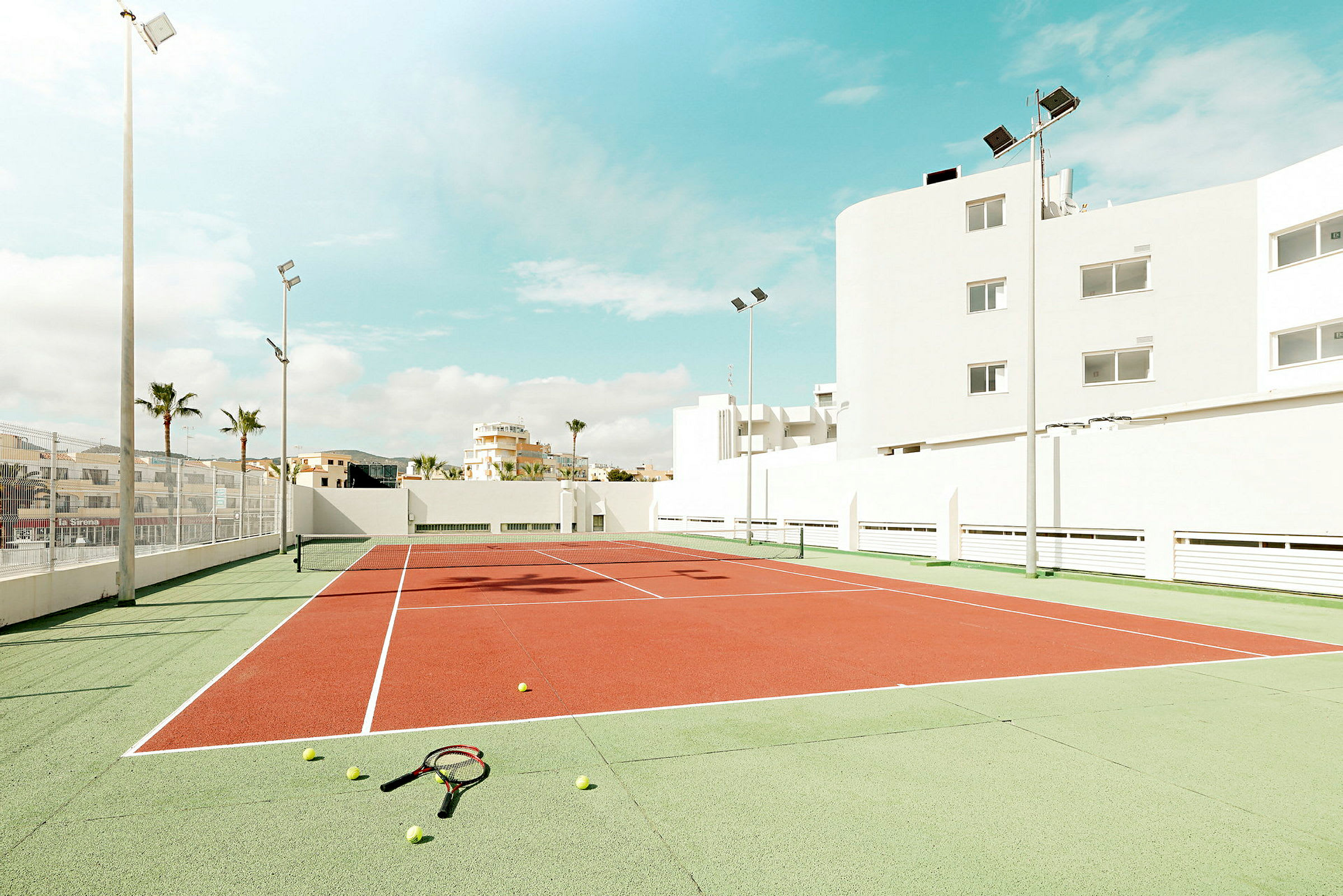 En tennisbane omgivet af bygninger under en solrig himmel, med tennisbolde og ketchere liggende på jorden. (AI-genereret tekst)