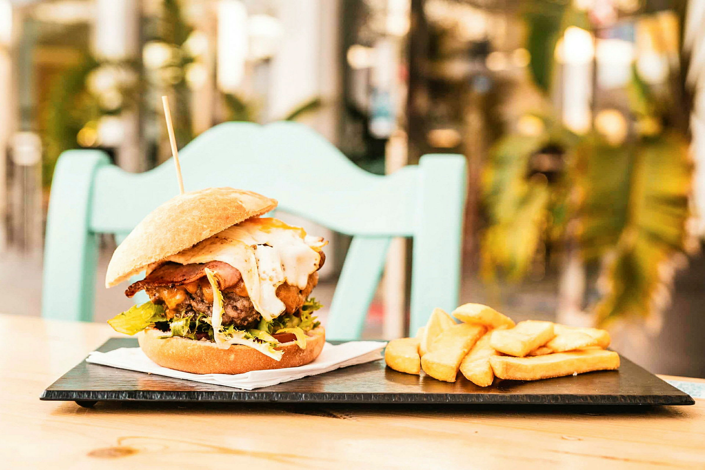 En burger med tilbehør af pommes frites på en tallerken, placeret på et bord med en stol i baggrunden. (AI-genereret tekst)