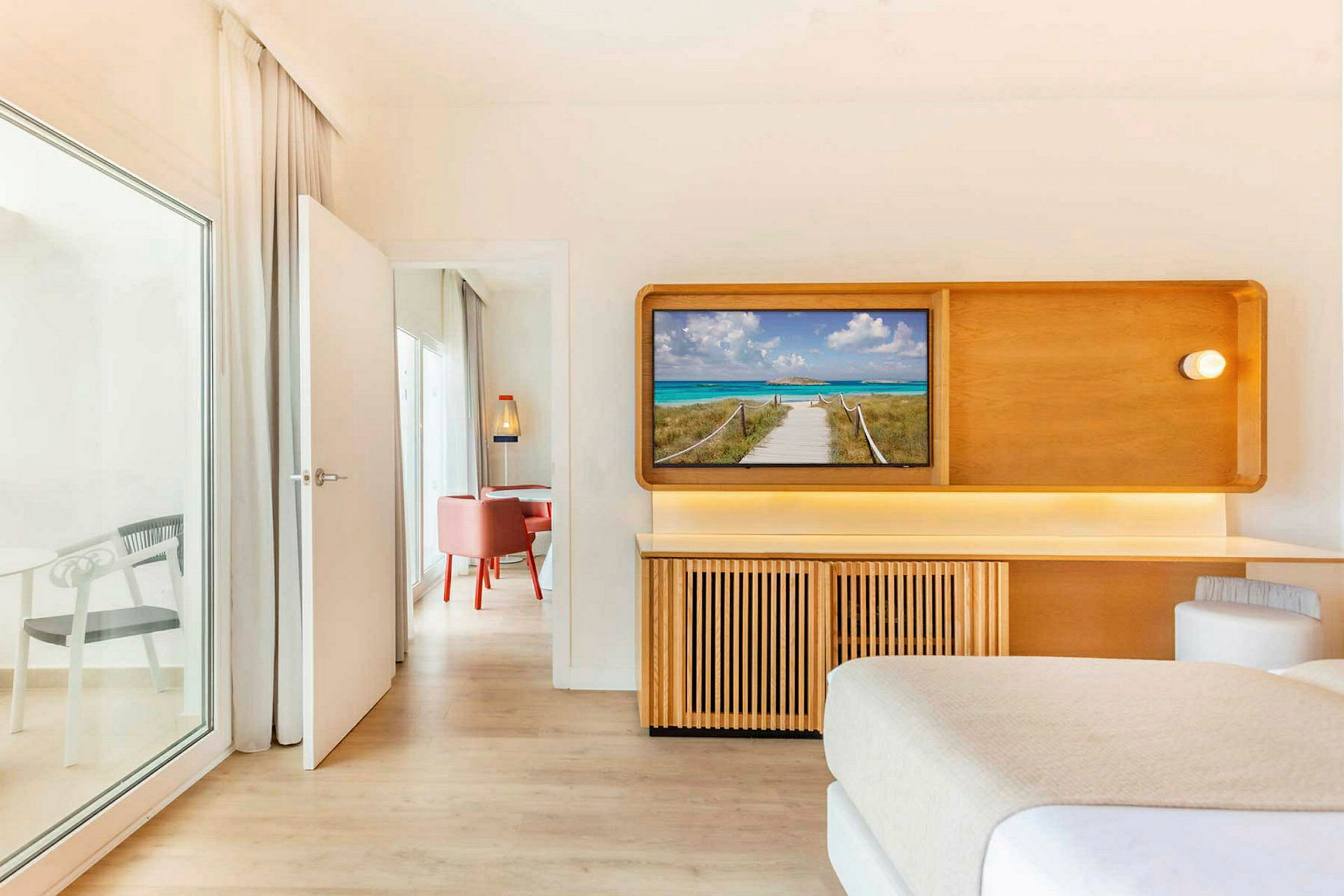 Et hotelværelse med et TV, der viser en strandpromenade. En dør fører ind til et tilstødende rum med røde stole. (AI-genereret tekst)