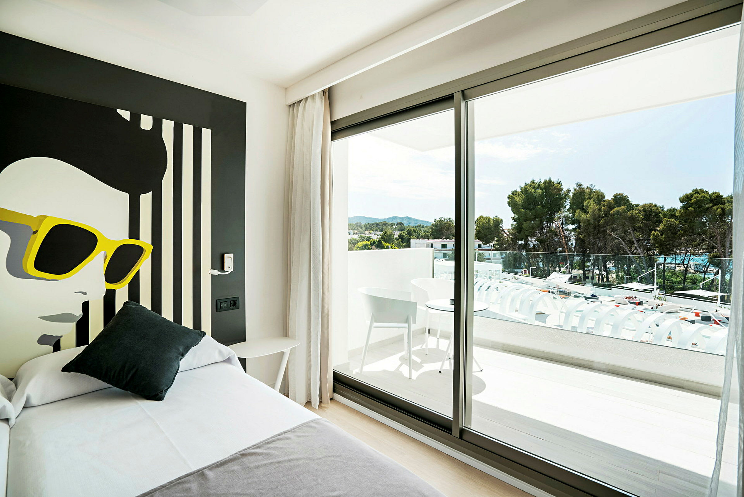 Et hotelværelse med moderne indretning, en seng i forgrunden og et stort vinduesparti, der fører ud til en balkon med møbler og udsigt over grønne træer. (AI-genereret tekst)