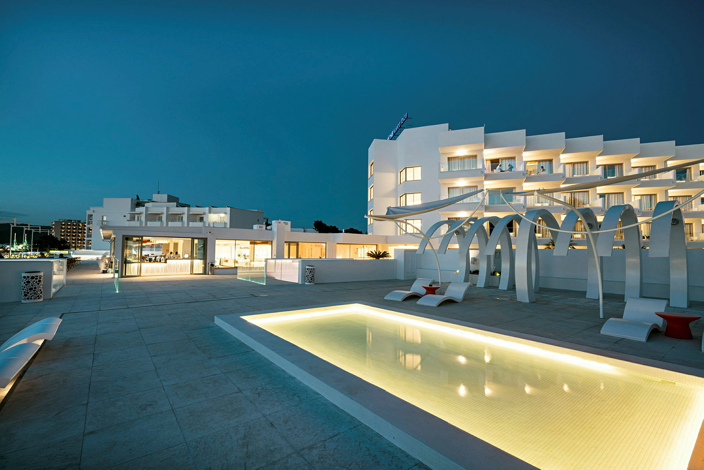 En moderne bygning med hvide facader, omgivet af en oplyst pool og en stilfuld terrasse. (AI-genereret tekst)