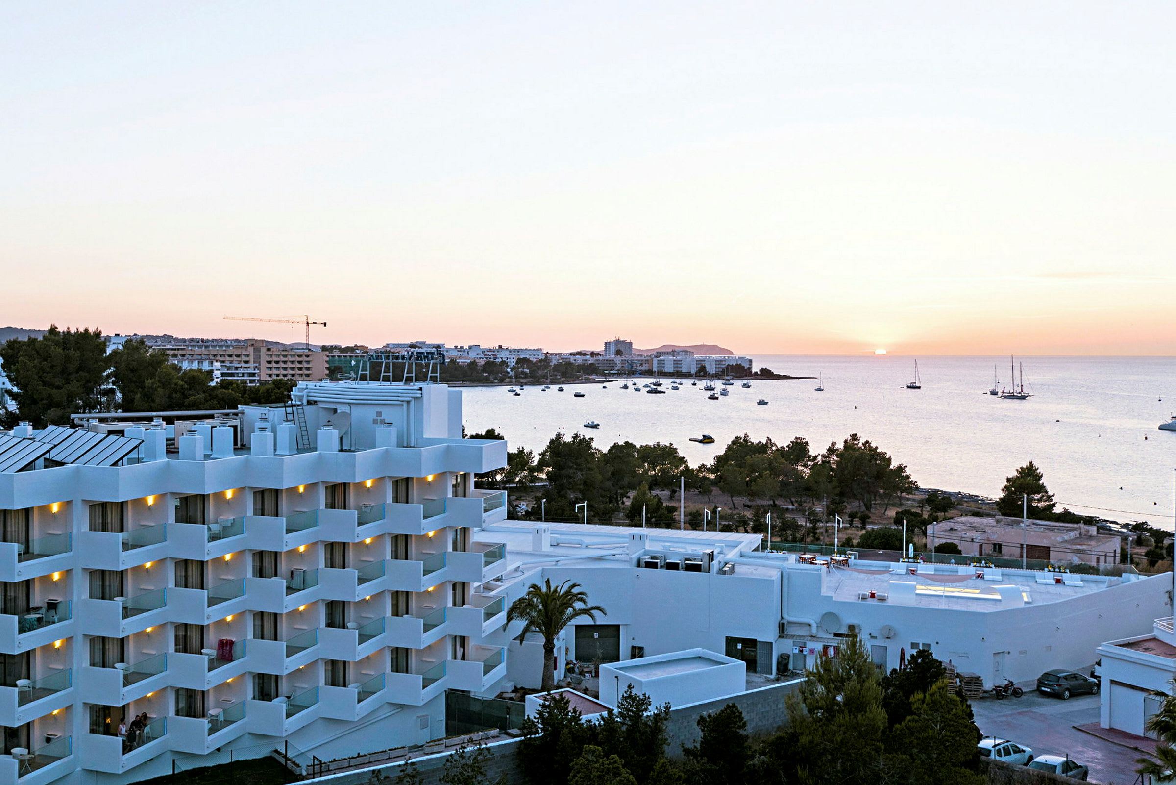 En udsigt over et hotel med hvide facader og altaner, omgivet af grønt, med havet og en solnedgang i baggrunden. (AI-genereret tekst)