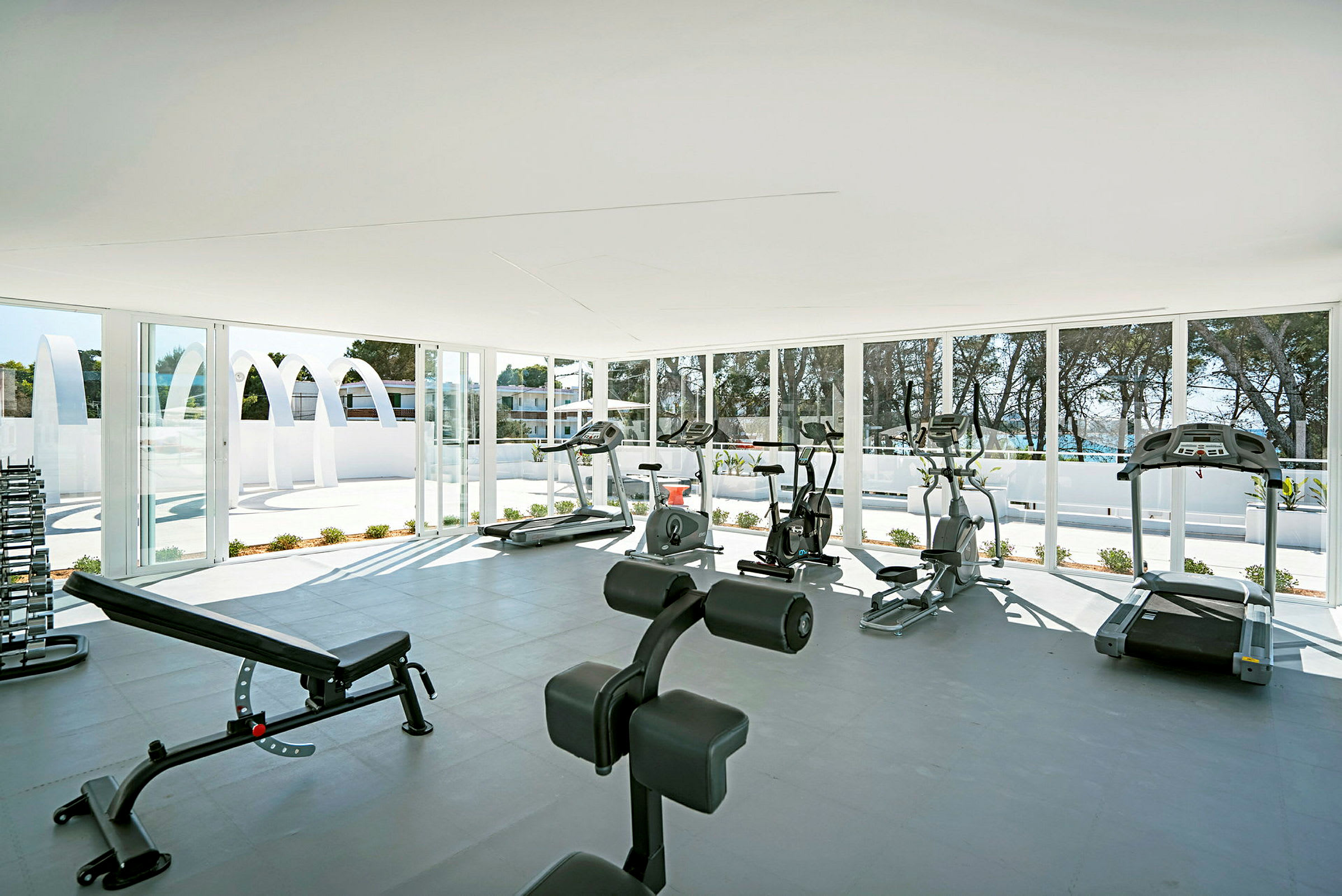 Et lyst og rummeligt fitnesscenter med forskellige træningsmaskiner og vægte, omgivet af vinduer med udsigt til udendørs omgivelser. (AI-genereret tekst)