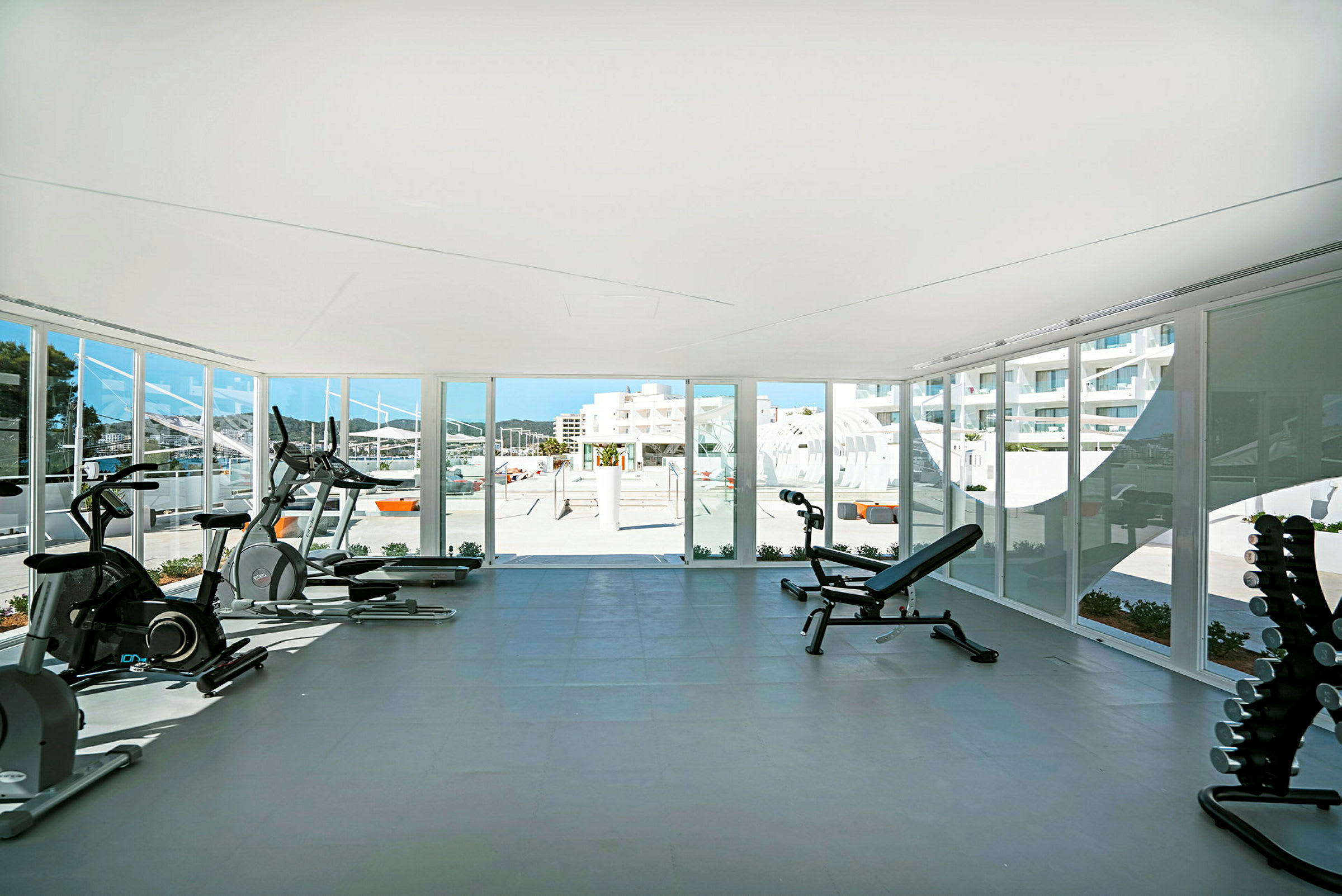 Fitnessområde med træningsudstyr og udsigt gennem store vinduer til et solrigt udendørsmiljø. (AI-genereret tekst)