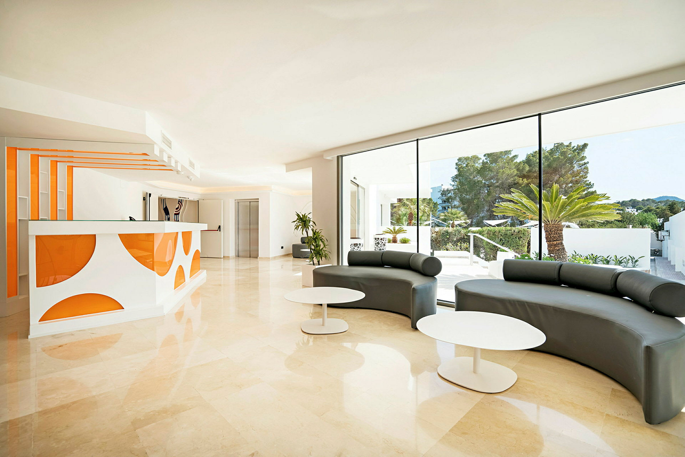 Lys hotel lobby med en receptionsskranke i hvid og orange samt siddeområder med moderne møbler. Store glasvinduer tillader naturligt lys og viser udendørsmiljøet. (AI-genereret tekst)