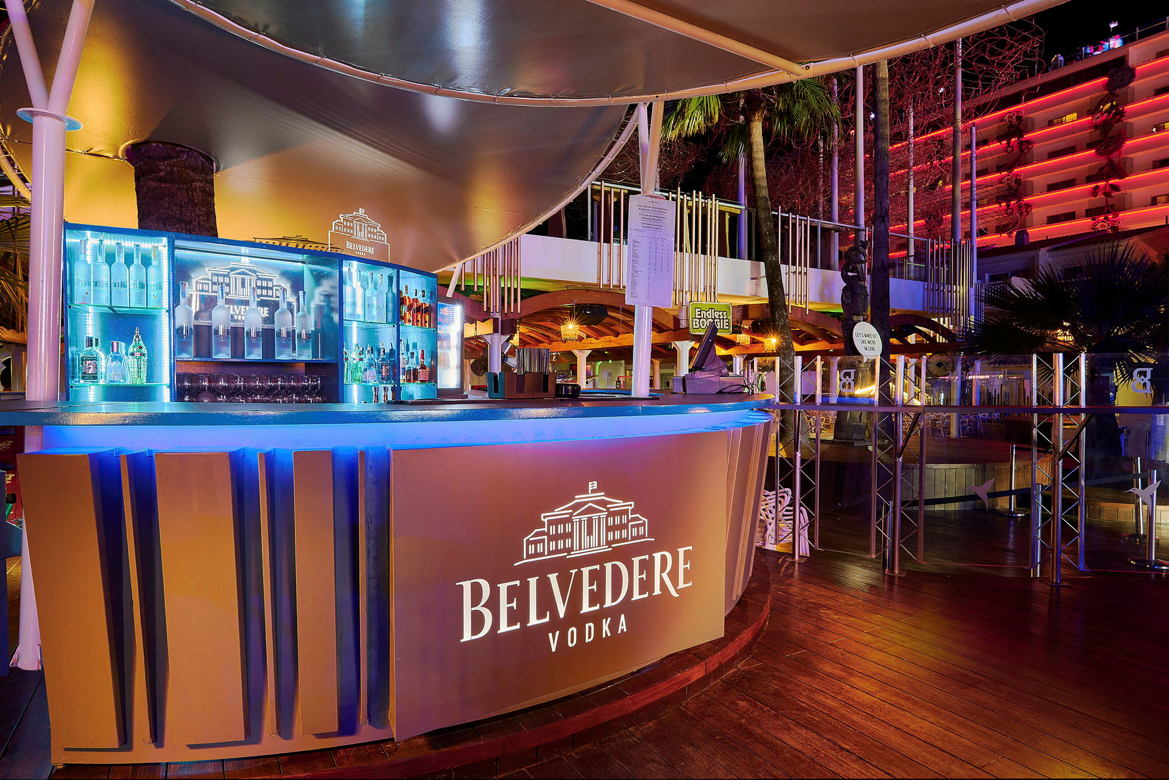 En udendørs bar med skilt for Belvedere Vodka, oplyst og med flasker i baggrunden. (AI-genereret tekst)
