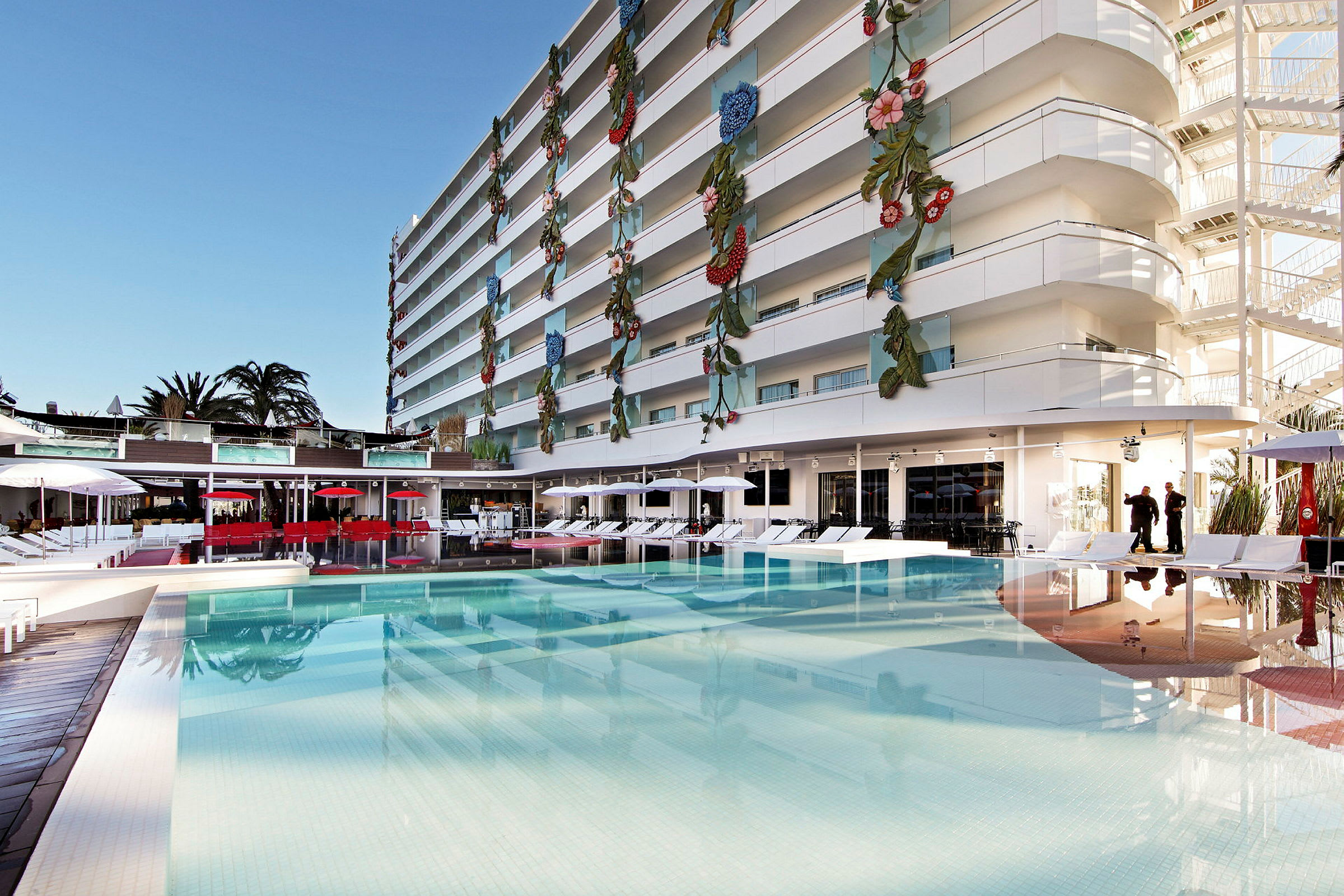 En moderne bygning med farvede dekorationer på facaden ses ved siden af en facilitet med en swimmingpool, liggestole og parasoller. (AI-genereret tekst)