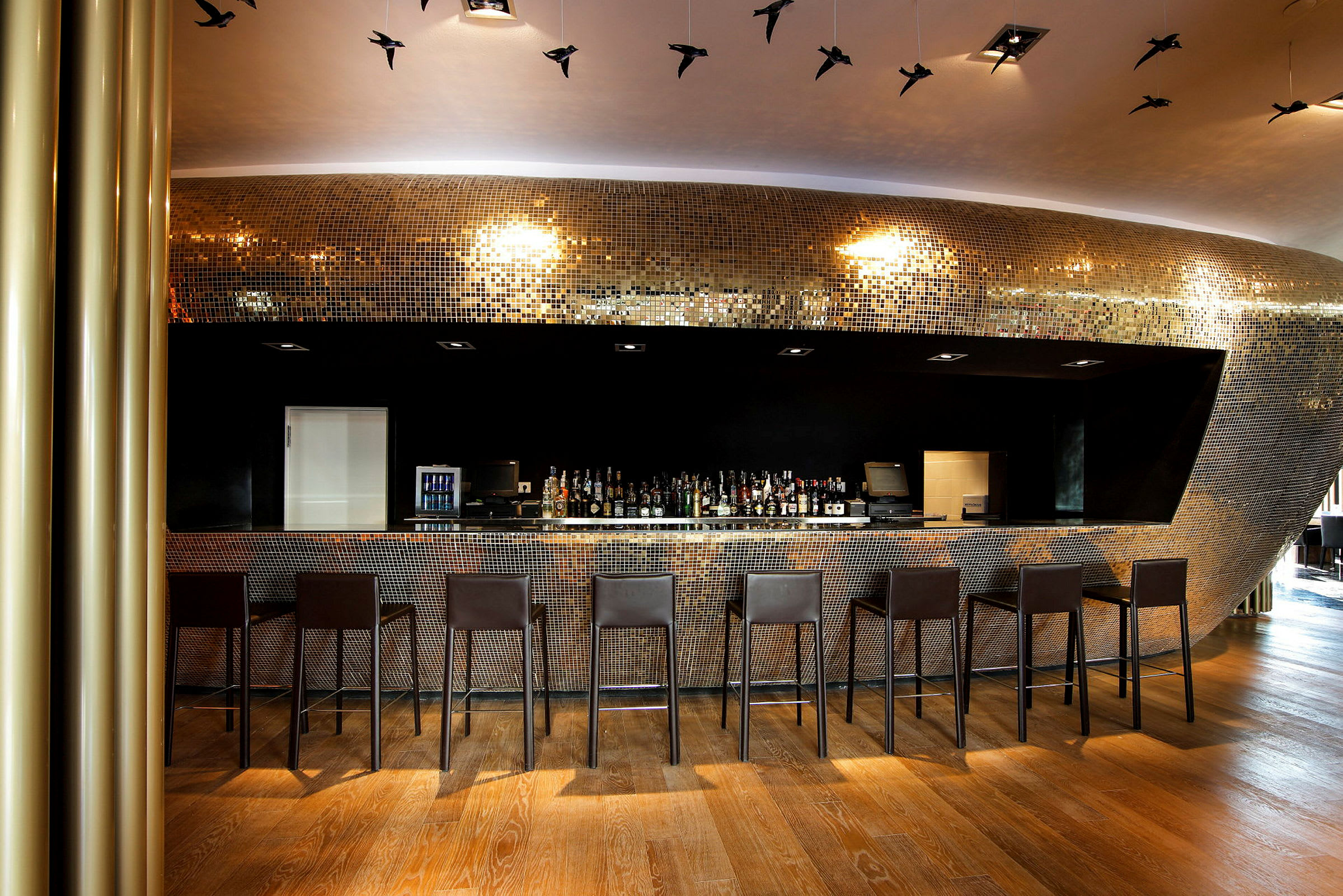 En moderne bar med høje stole foran en disk dækket af skinnende mosaik. Loftet har dekorative fugle i luften. (AI-genereret tekst)