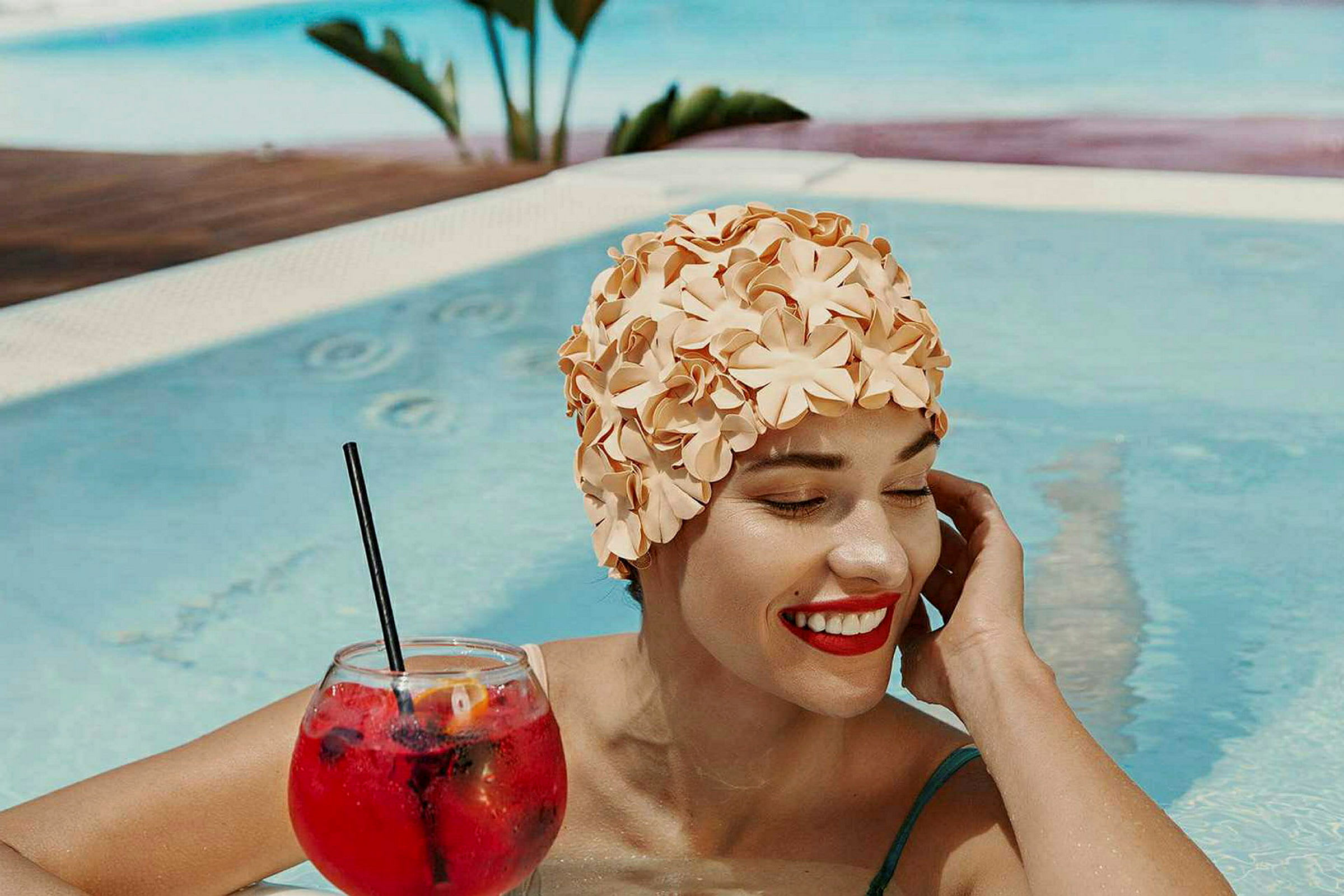 En person i en pool har en blomsteret badehætte på og holder et glas med en rød drink. (AI-genereret tekst)