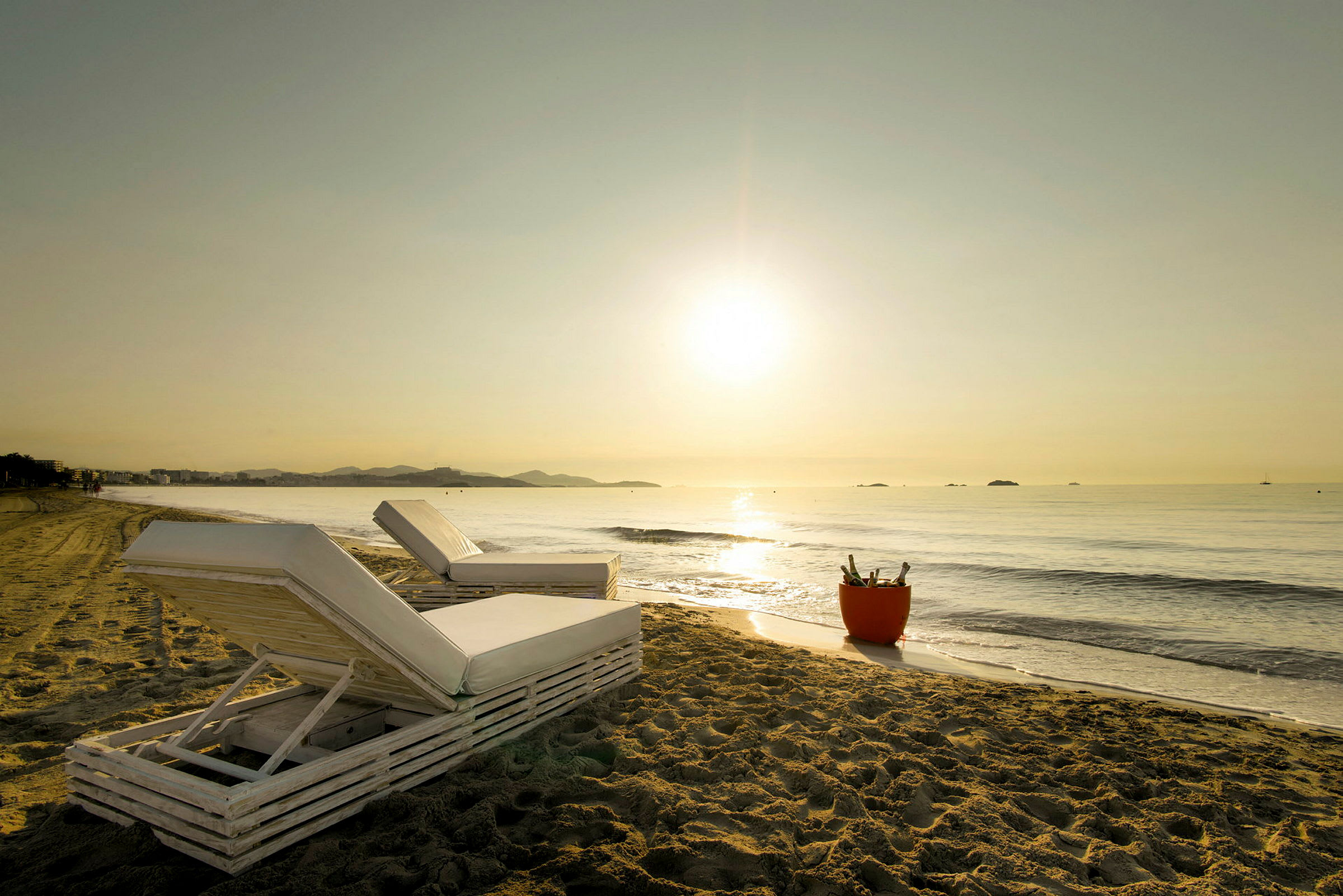 Solstole på en sandstrand med udsigt over havet ved solopgang eller solnedgang. En spand med flasker står ved siden af. (AI-genereret tekst)