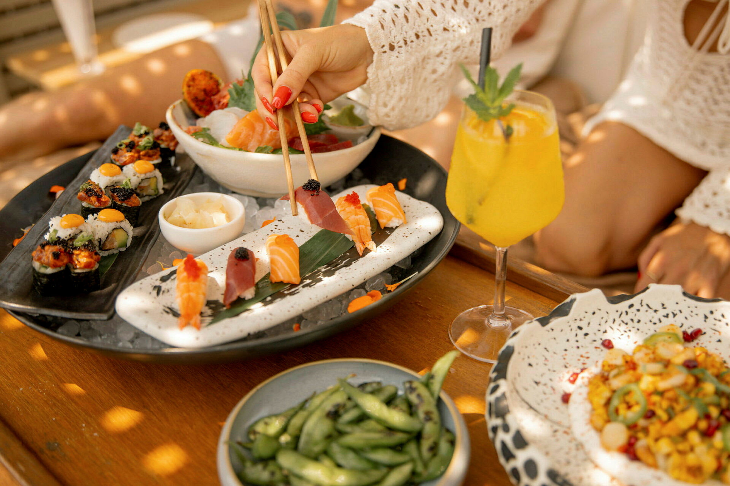 En tallerken med sushi og sashimi sammen med en gul drik og en skål sojabønner, placeret på et træbord. (AI-genereret tekst)