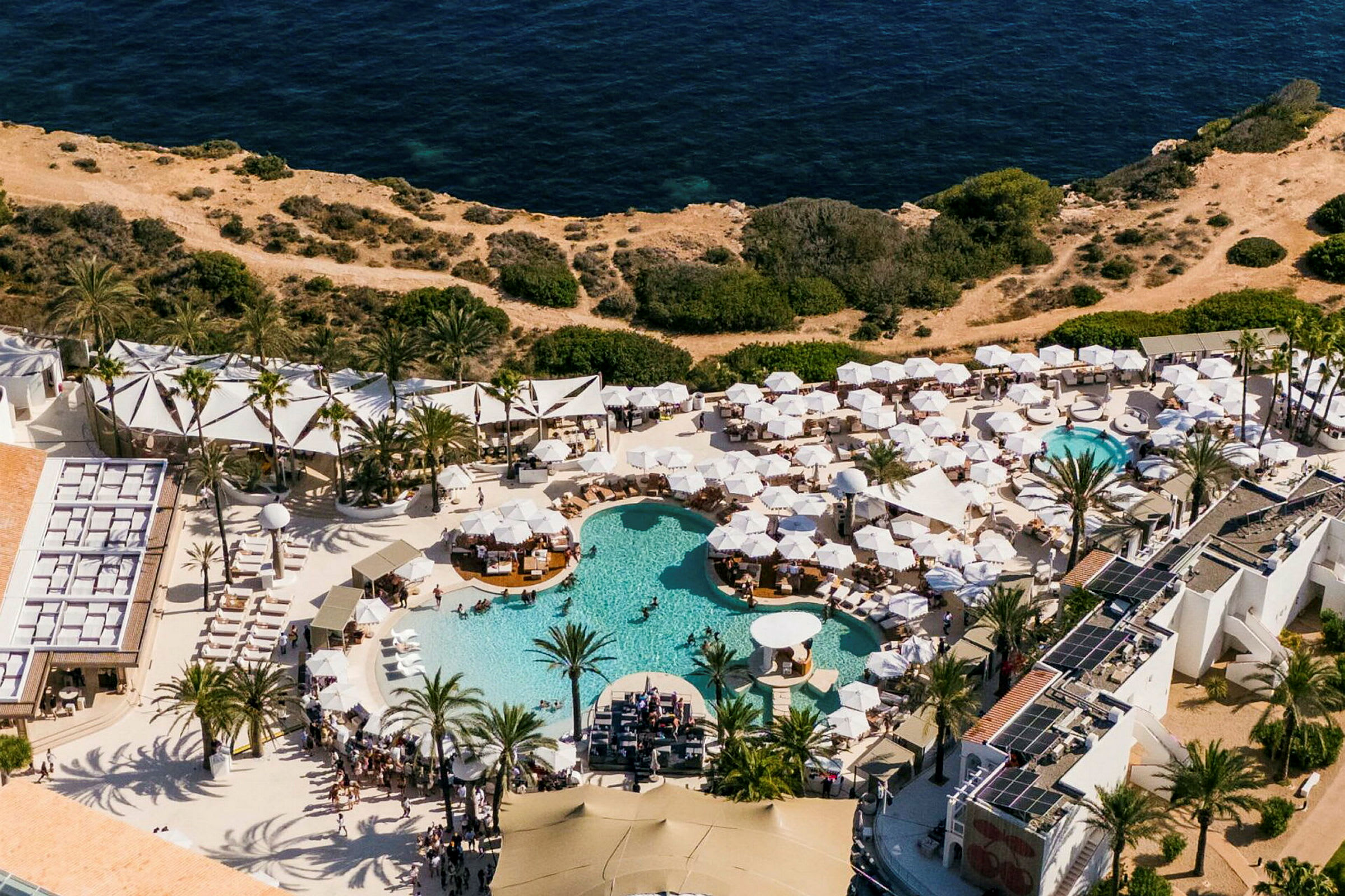 Luftfoto af en ferieresort med flere pools og hvide parasoller, ved siden af klipper og havet. (AI-genereret tekst)