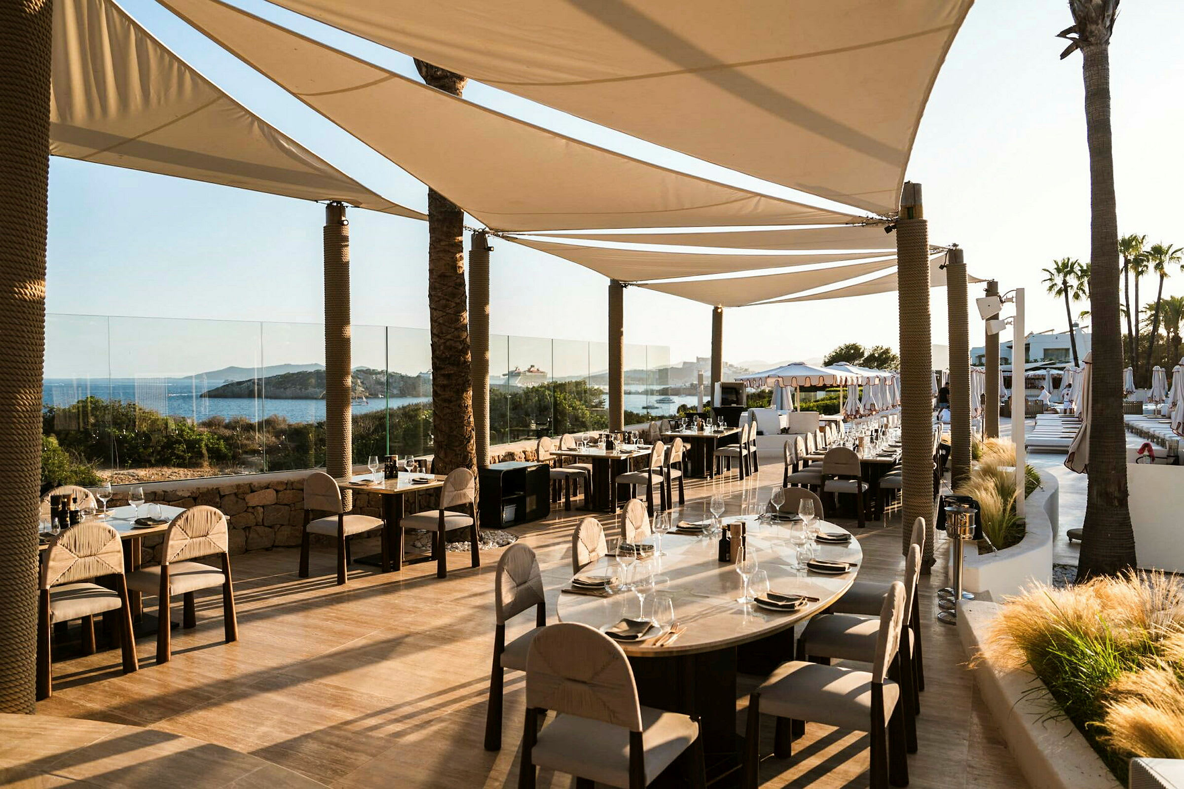 Restaurant med borde og stole på en terrasse tæt på havet, skygget af sejldug. (AI-genereret tekst)