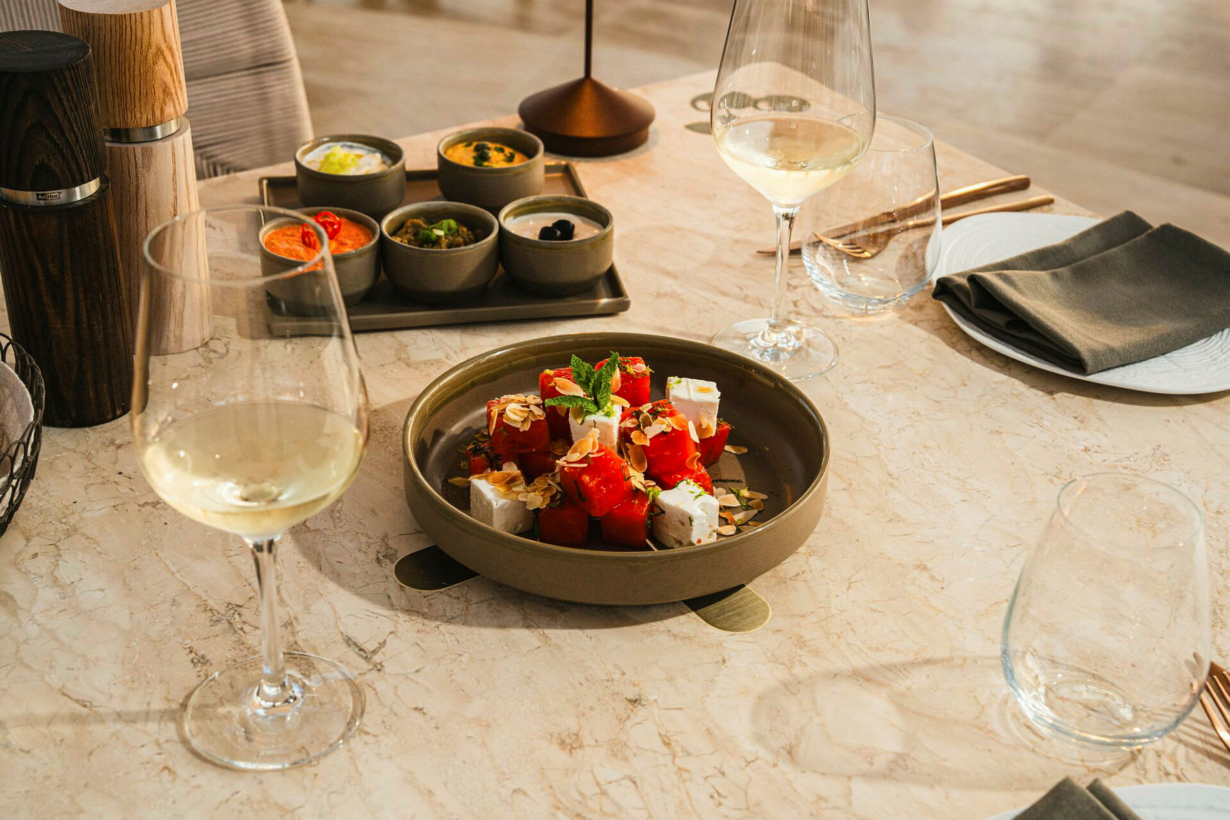 Et bord med en tallerken med salat indeholdende vandmelon og fetaost omgivet af forskellige tilbehør i små skåle samt glas med hvidvin. (AI-genereret tekst)