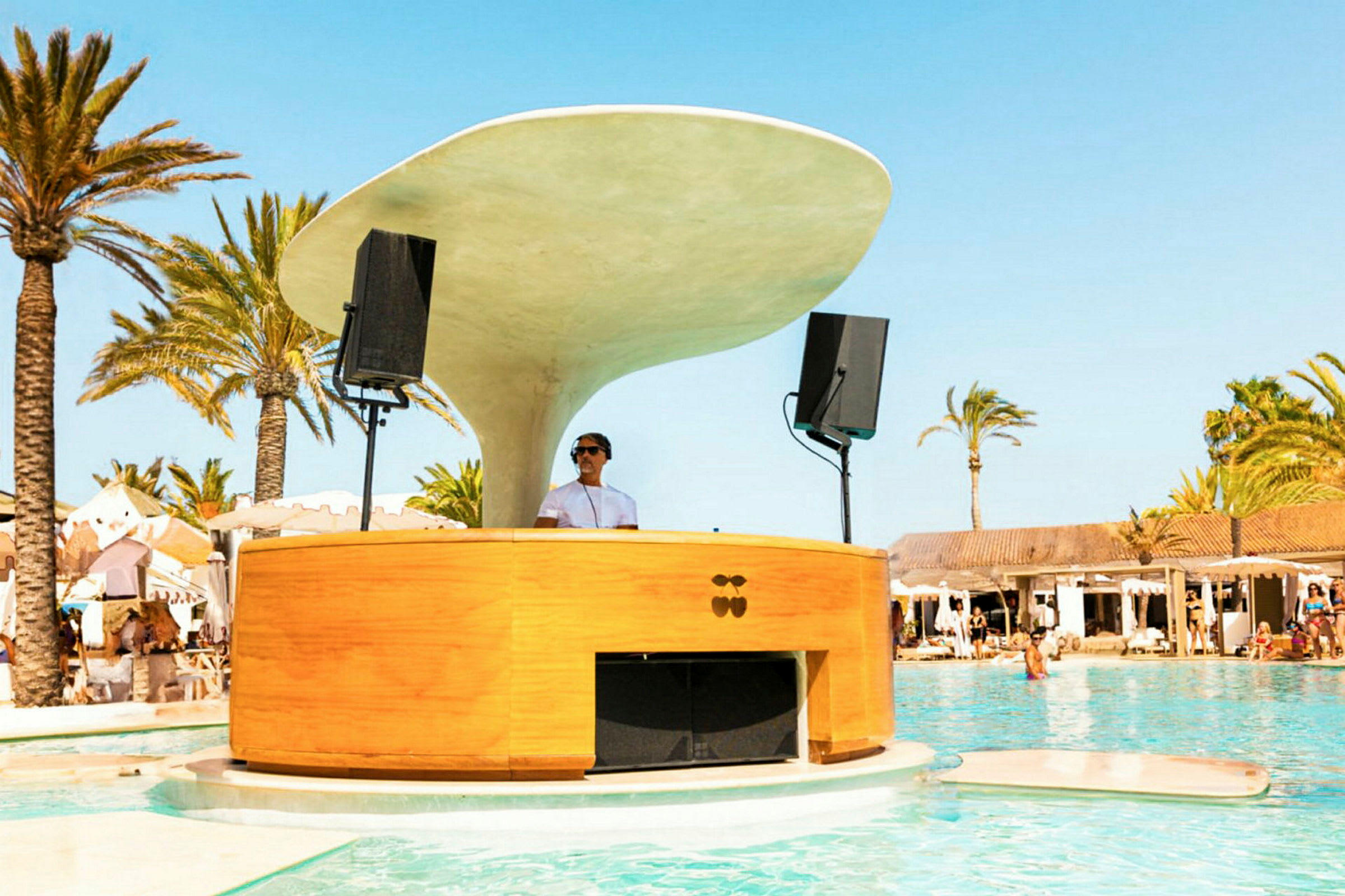 En DJ-platform ved en pool i en strandnær atmosfære med palmer og bygninger i baggrunden. (AI-genereret tekst)