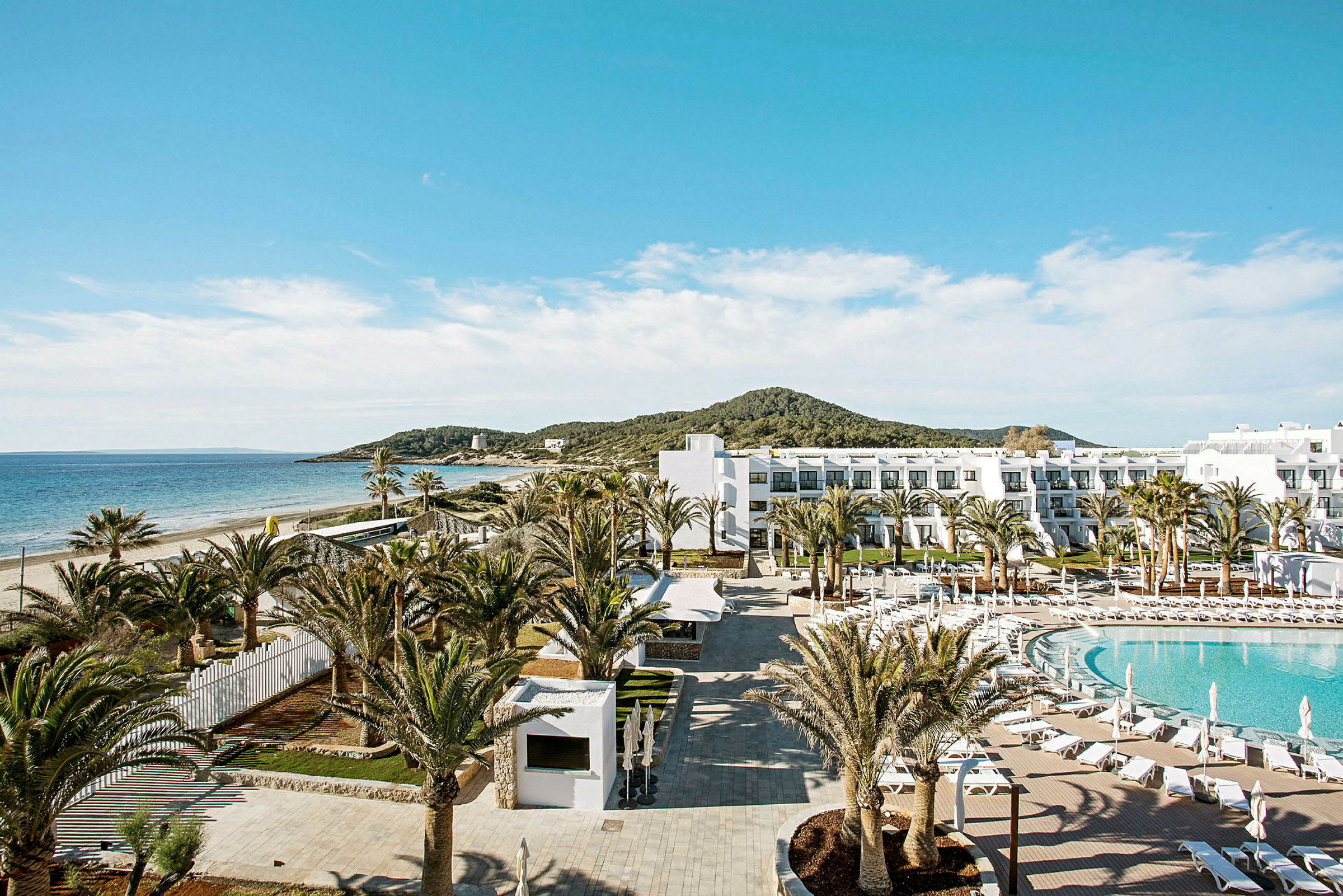 Et hotelområde ved stranden med pool, palmer og en hvid bygning. (AI-genereret tekst)