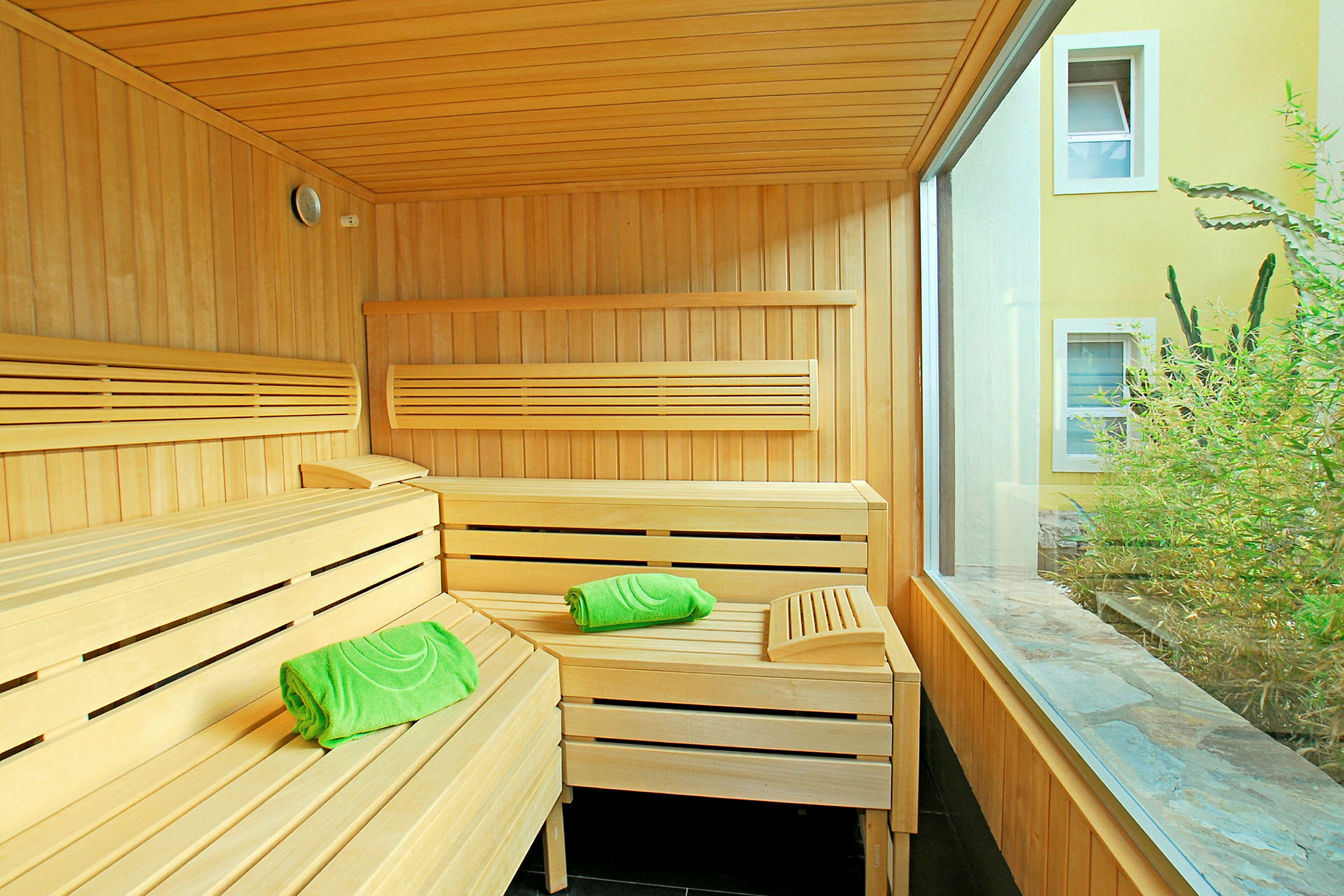 En sauna i træ med grønne håndklæder og store vinduer mod en gårdhave. (AI-genereret tekst)