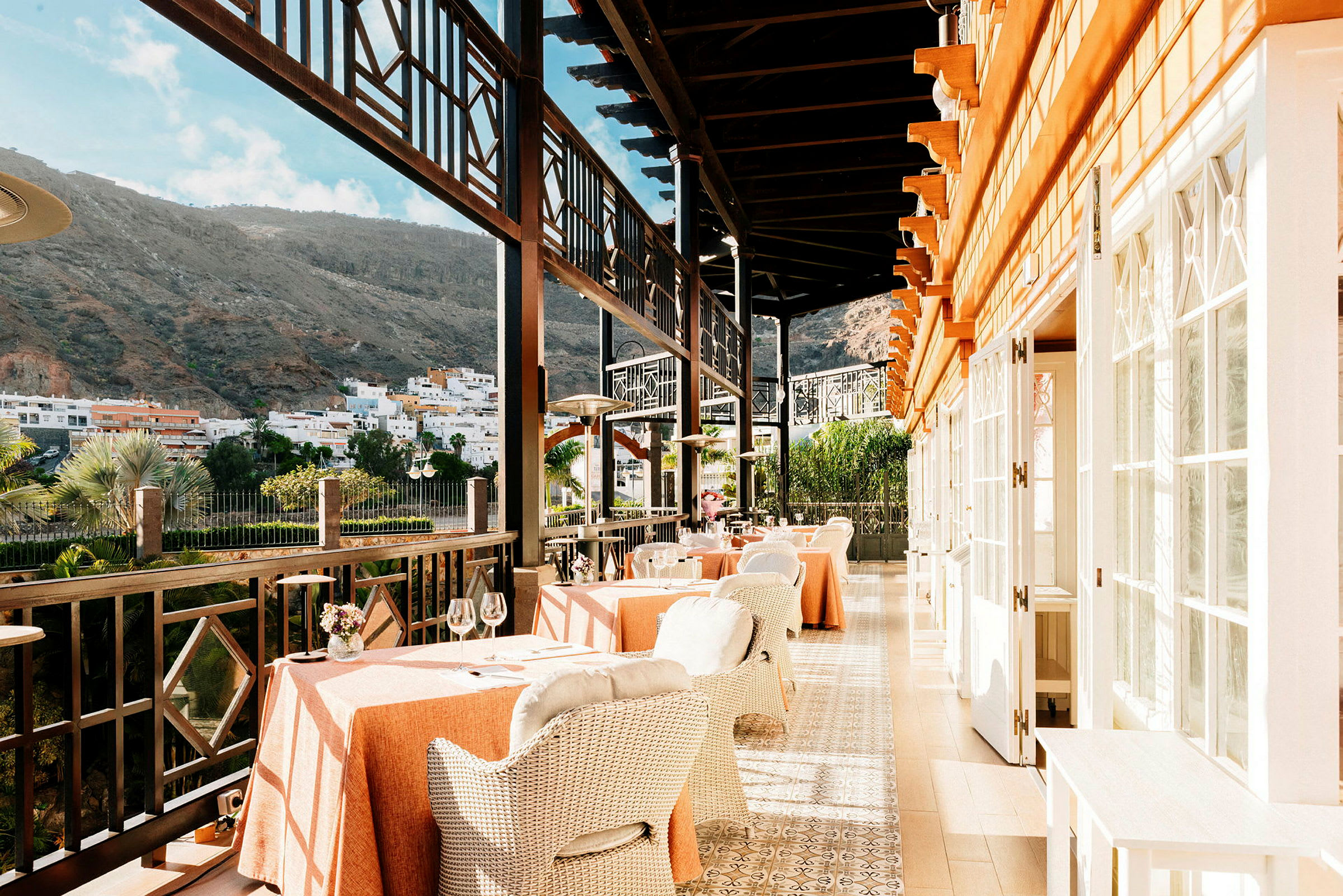 Restaurant med borde og stole på en terrasse med udsigt over bygninger og bjerge. (AI-genereret tekst)