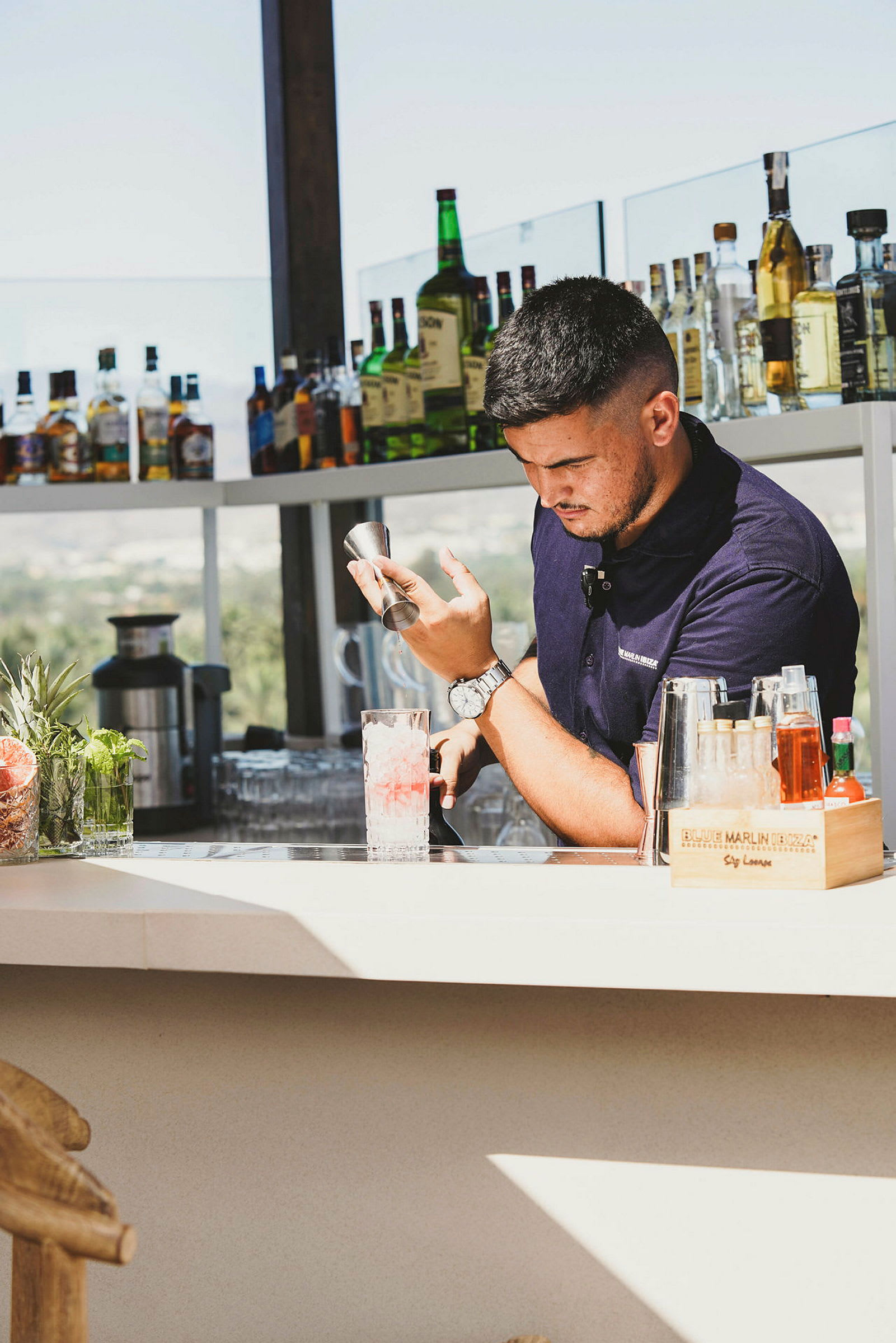En bartender ved en bar med flasker og drikkevarer på hylder. (AI-genereret tekst)