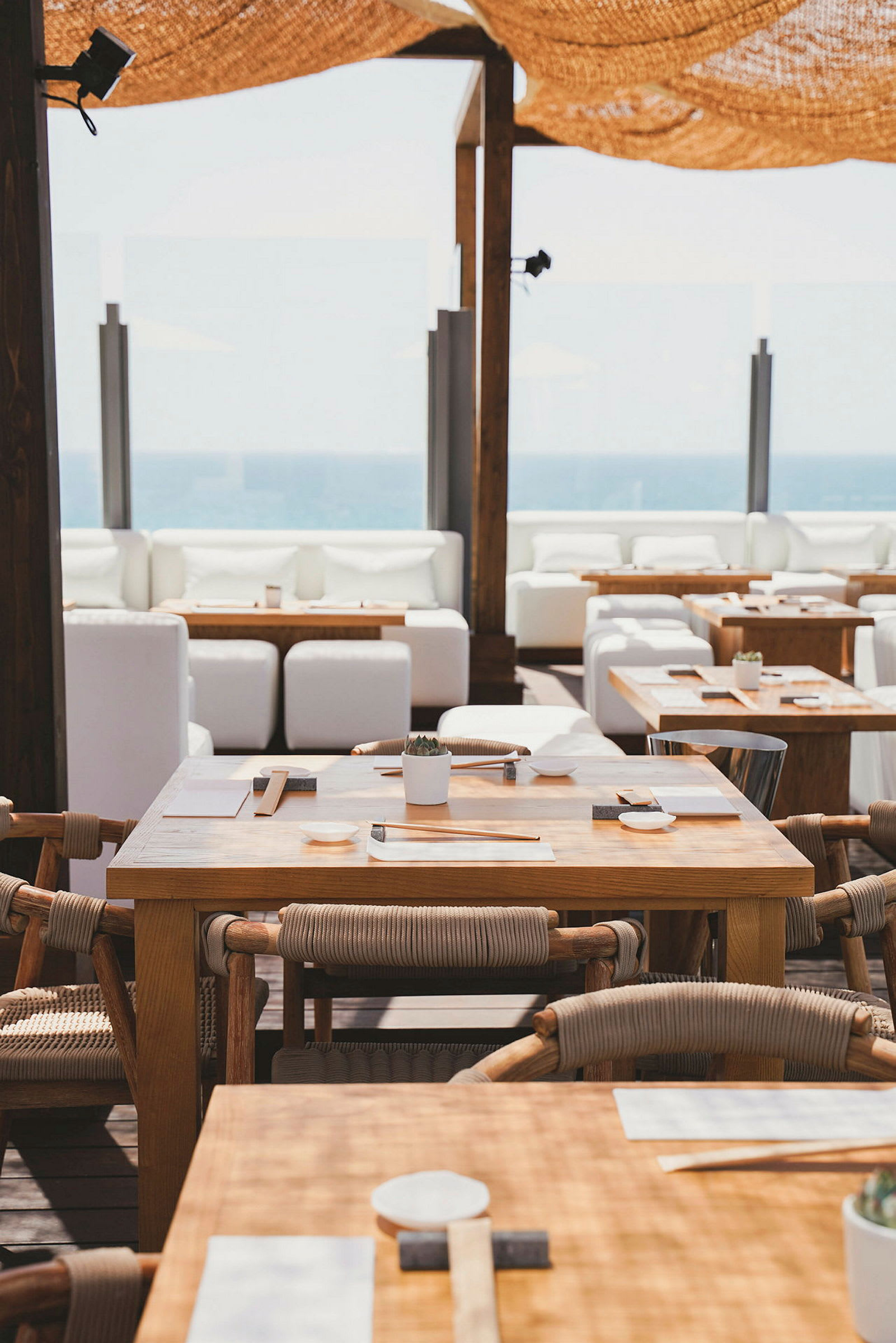 Udendørsrestaurant med træborde og stole, hvide sofaer i baggrunden samt udsigt over havet. (AI-genereret tekst)