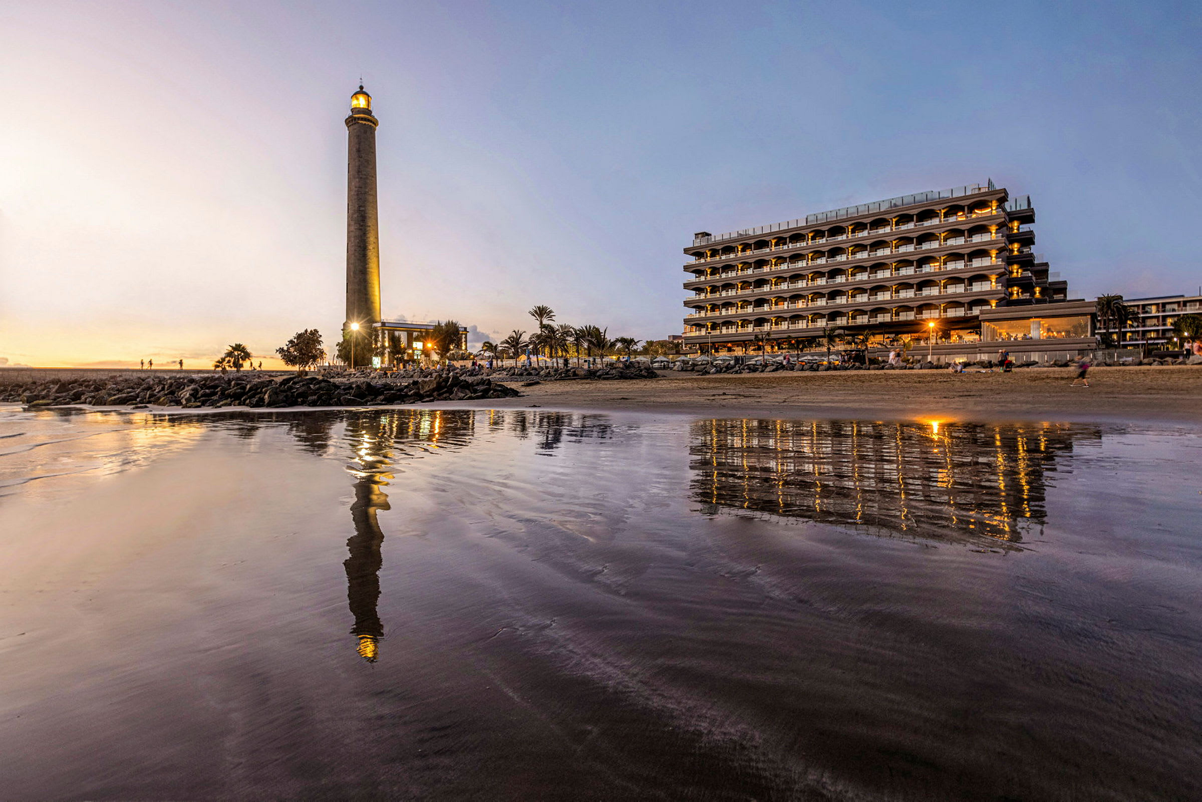 Billede af et fyr ved stranden med et hotel i baggrunden, refleksioner synlige i vandoverfladen. (AI-genereret tekst)