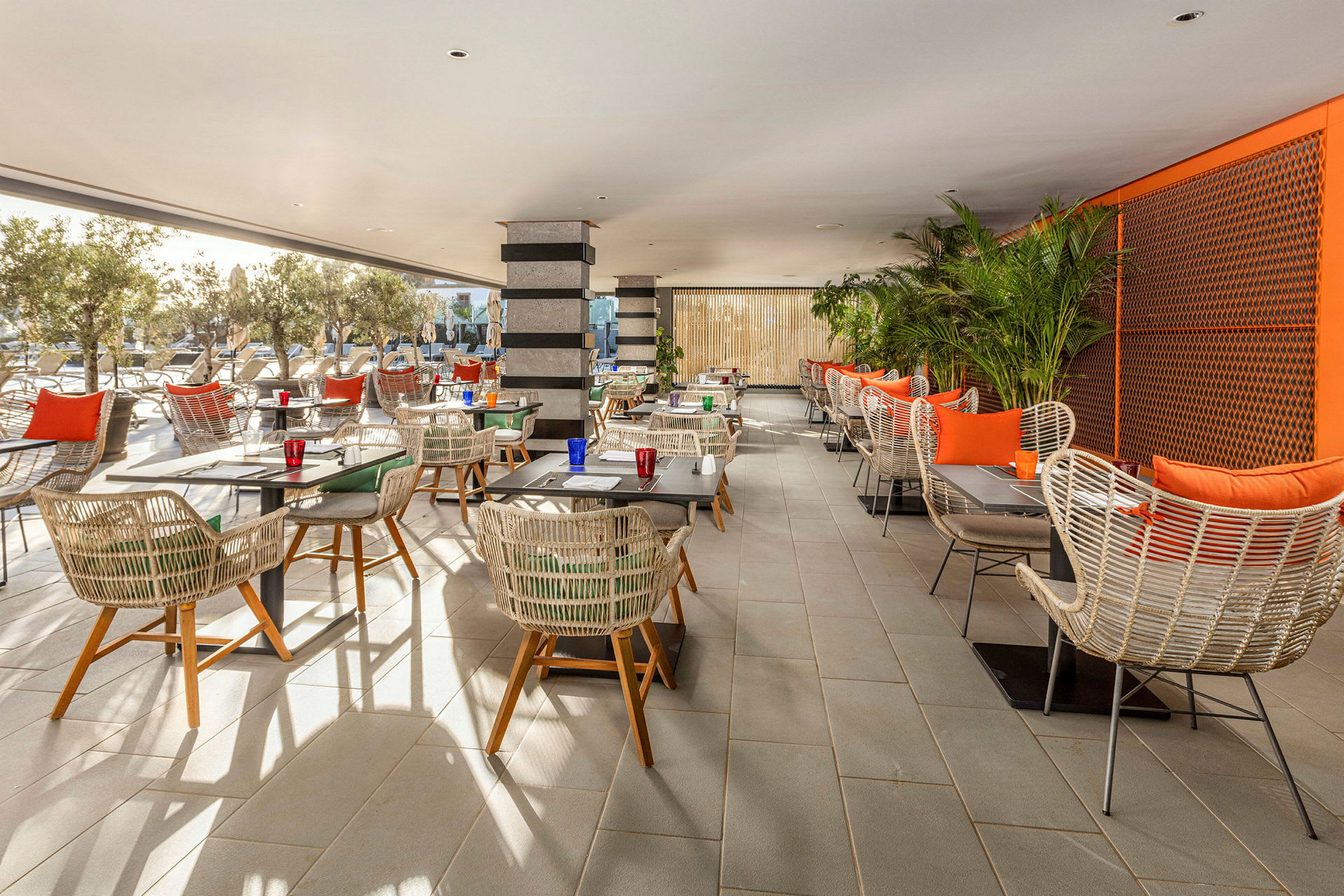 En restaurant med borde og stole i rottingstil, omgivet af planter og udsigt til et udendørsområde. (AI-genereret tekst)