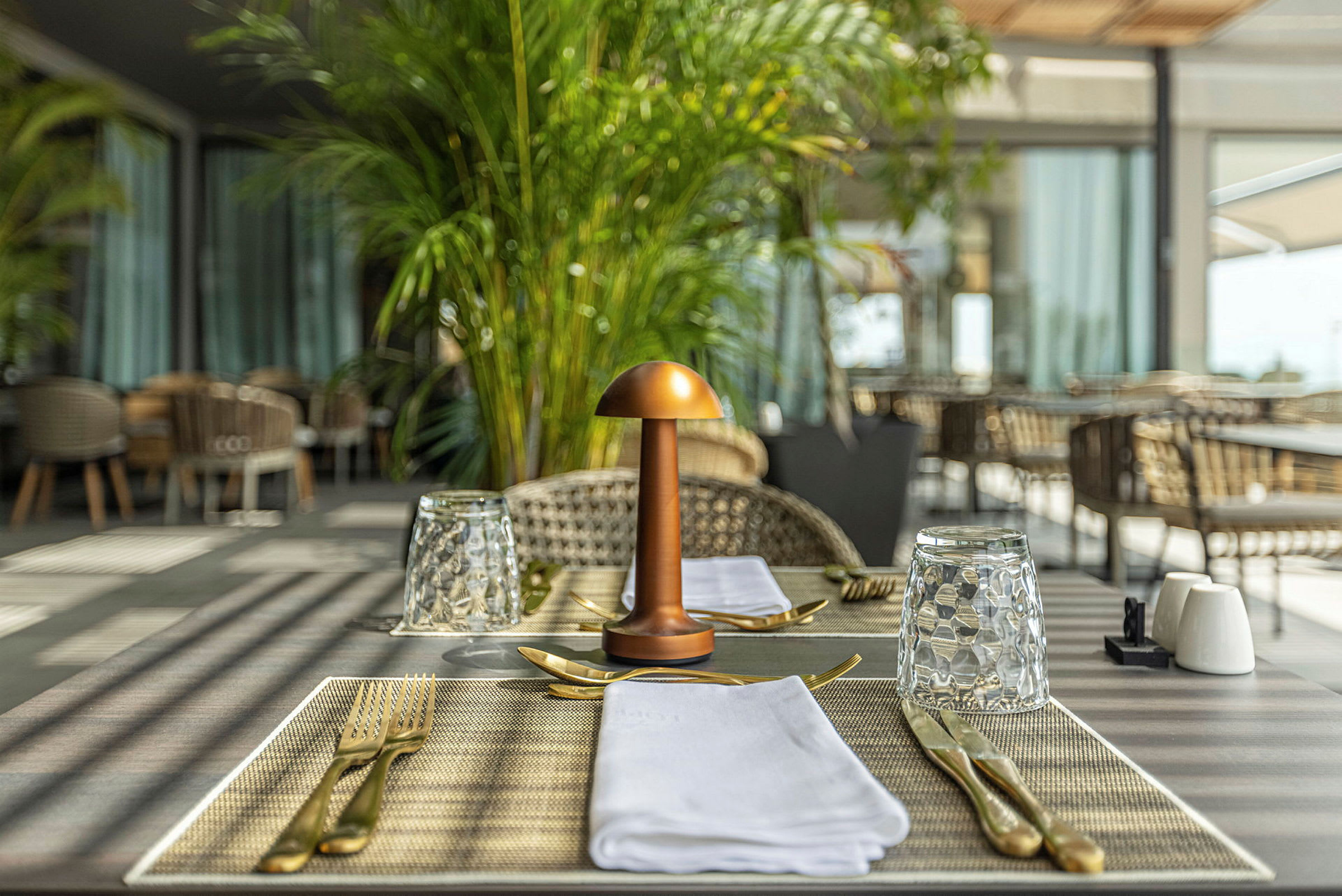 Et bord i et restaurantområde er dækket med glas, bestik og servietter. En dekorativ lampe er placeret i midten. (AI-genereret tekst)