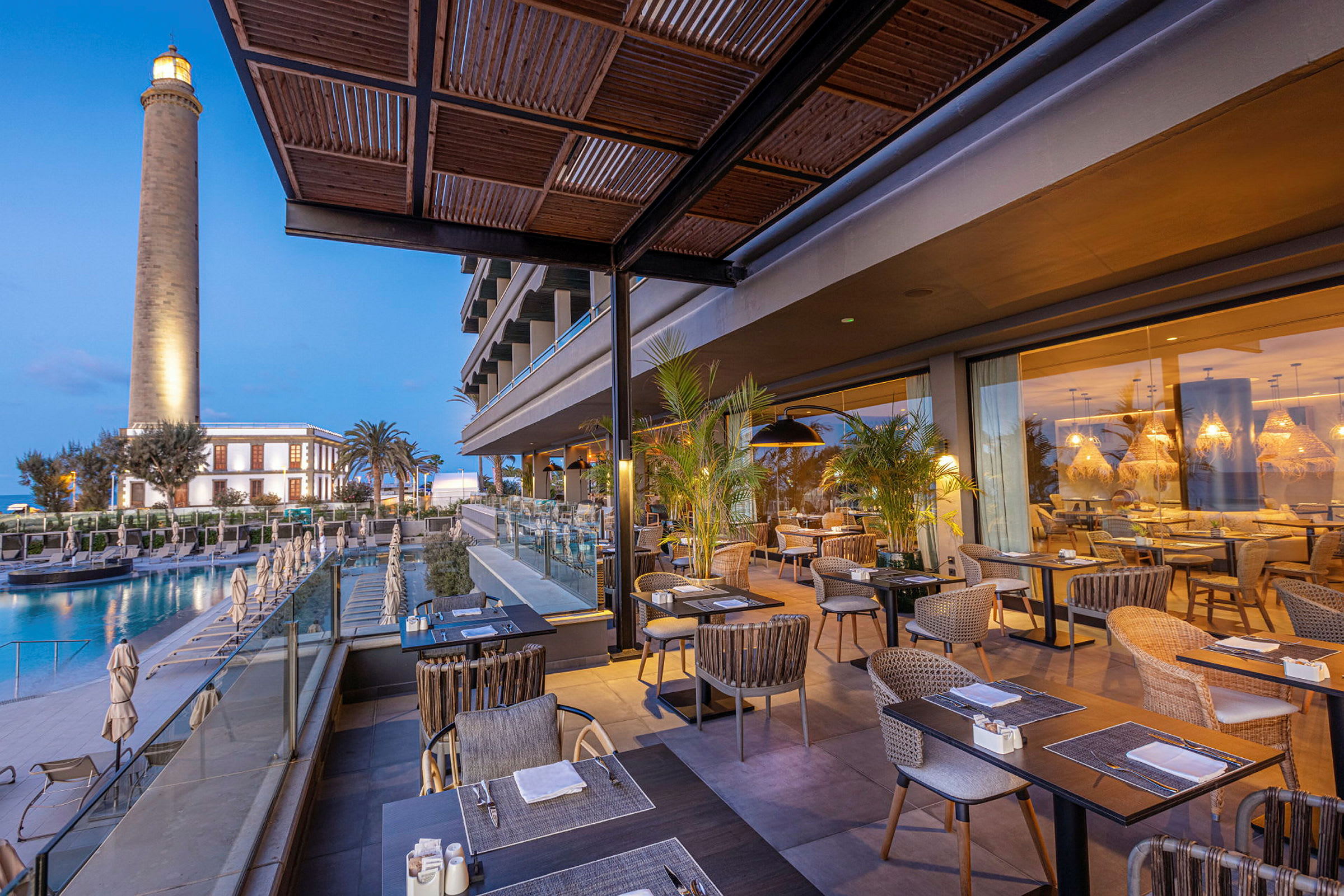Restaurant med udendørsborde ved et hotel, et fyrtårn i baggrunden. (AI-genereret tekst)