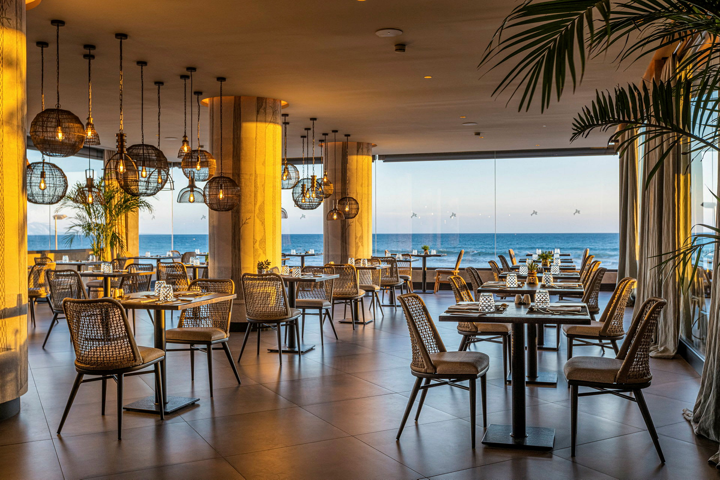 Billede af en restaurant med kurvestole, bordene er dækket med udsigt over havet gennem store glasvinduer. (AI-genereret tekst)