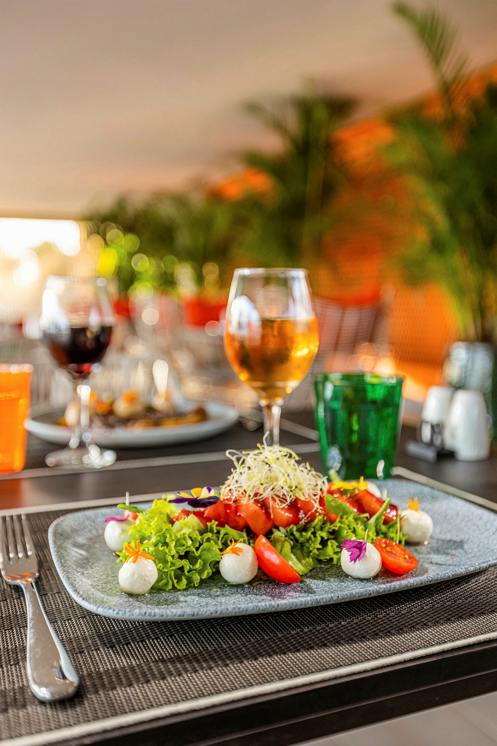 En tallerken med farverig salat, tomater og mozzarella-kugler står på et dækket bord. Baggrunden viser et glas med drikkelse og grønne planter. (AI-genereret tekst)