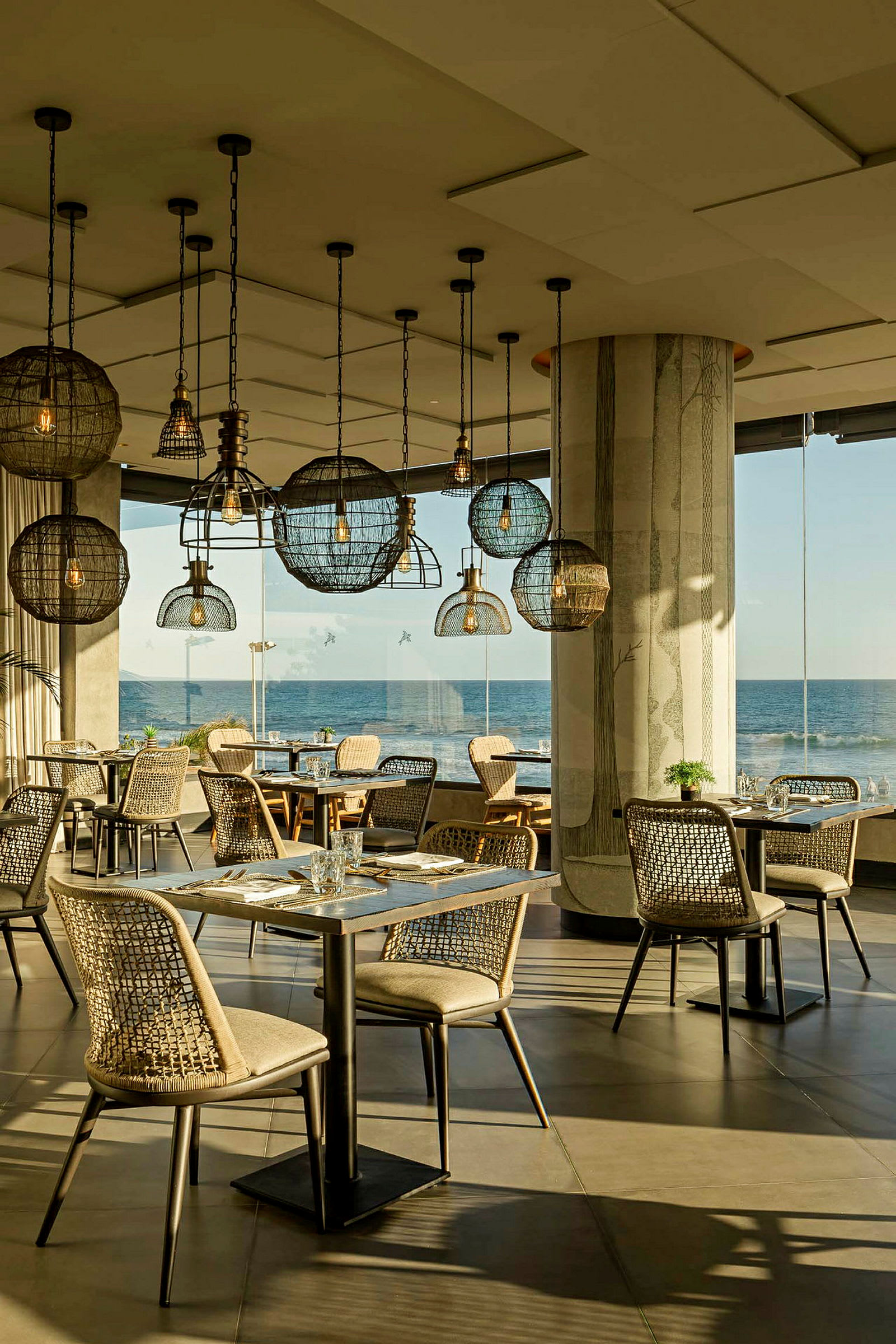 En restaurant med moderne indretning, flere spiseborde og stole samt dekorative lamper i loftet. Vinduer med udsigt over havet. (AI-genereret tekst)