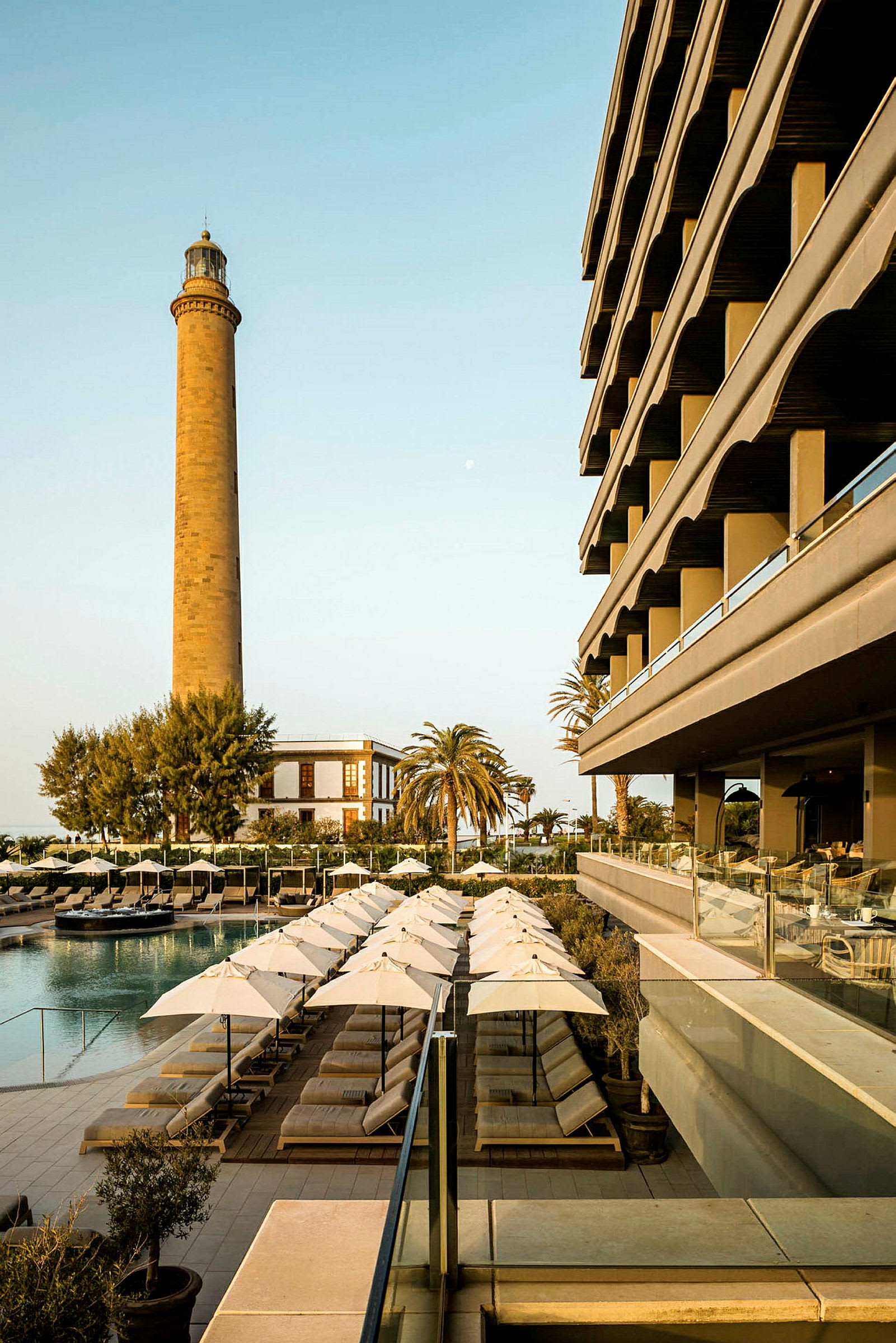 Billede af en hotelbygning ved siden af en pool med liggestole og parasoller. Et fyrtårn og en palme ses i baggrunden. (AI-genereret tekst)