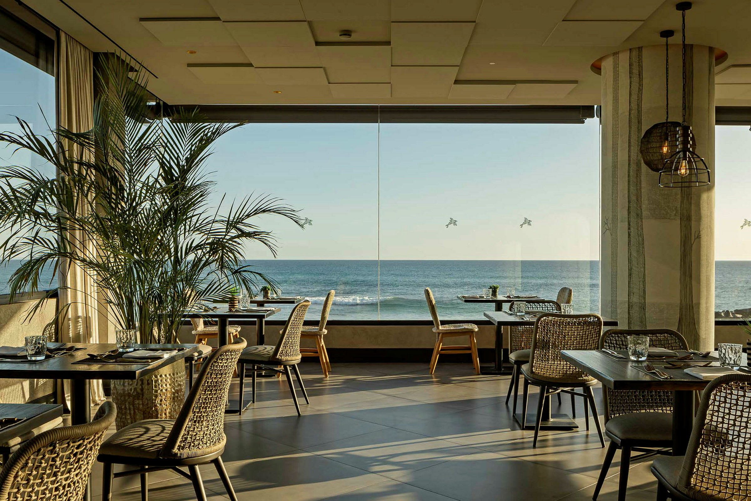 En lys restaurant med store vinduer med udsigt over havet. Borde og stole er pænt arrangeret, og grønne planter pryder interiøret. (AI-genereret tekst)