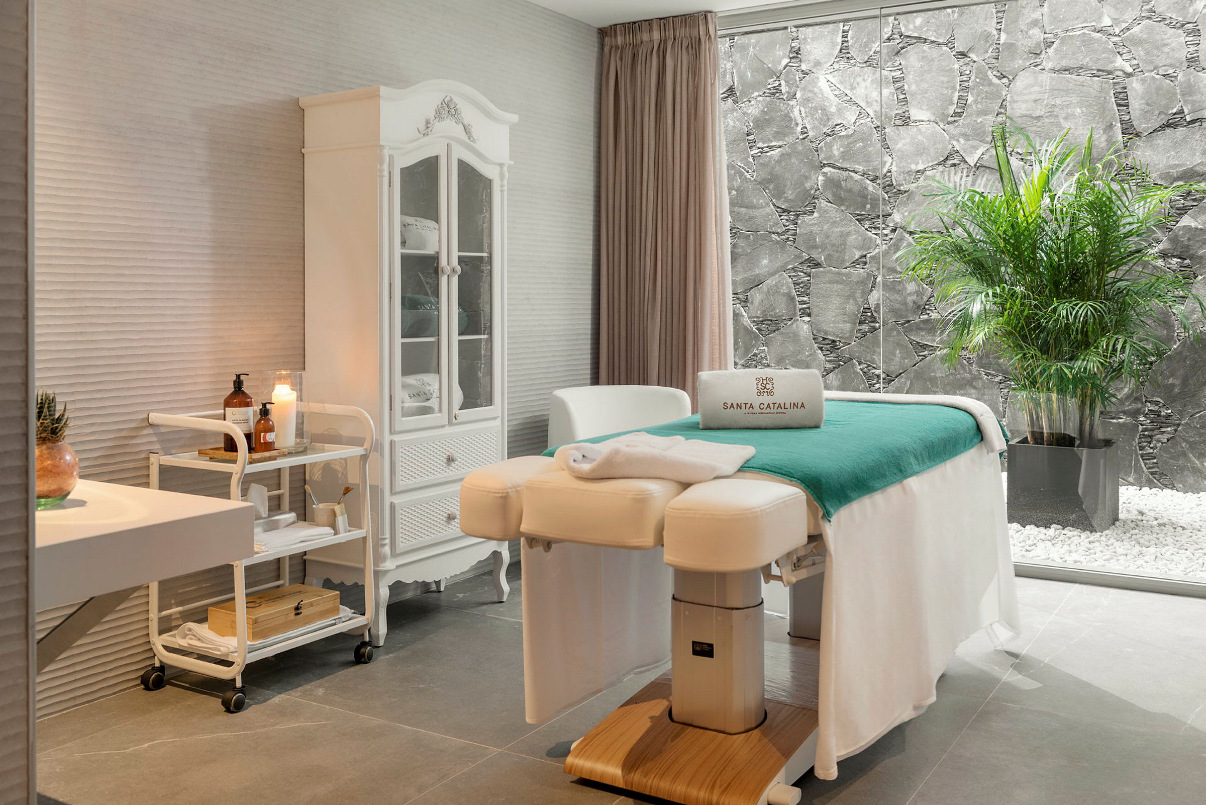Et stilfuldt spa-rum med massagebriks, hvid indretning og et stort vindue med udsigt til en stenmur og grønne planter. (AI-genereret tekst)