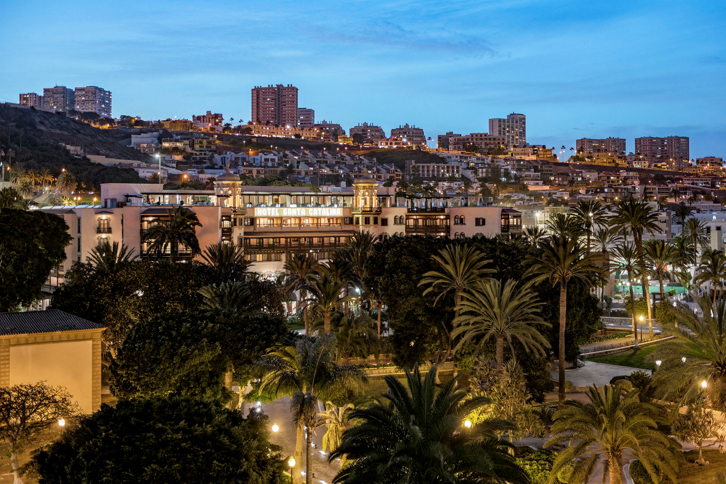 Hotel Santa Catalina omgivet af træer med en baggrund af byen om aftenen. (AI-genereret tekst)